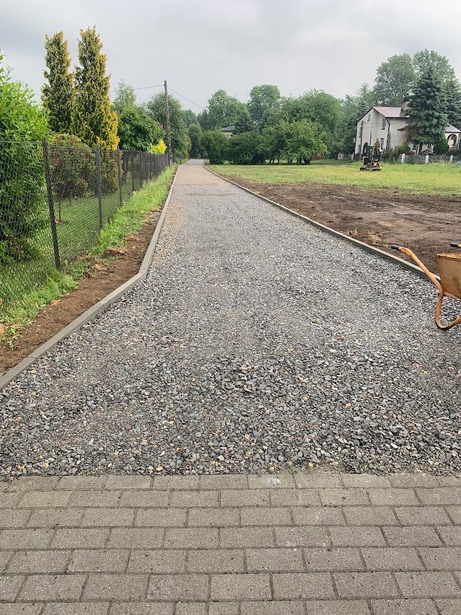 Świeżo utwardzona droga szutrowa z betonowymi obrzeżami, widok z perspektywy chodnika z kostki, w tle dom i zieleń.