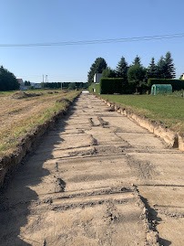Przygotowanie podłoża pod drogę gruntową, widoczne wyrównane i utwardzone fragmenty ziemi, po bokach wykopane rowy, w tle zabudowa jednorodzinna i drzewa.