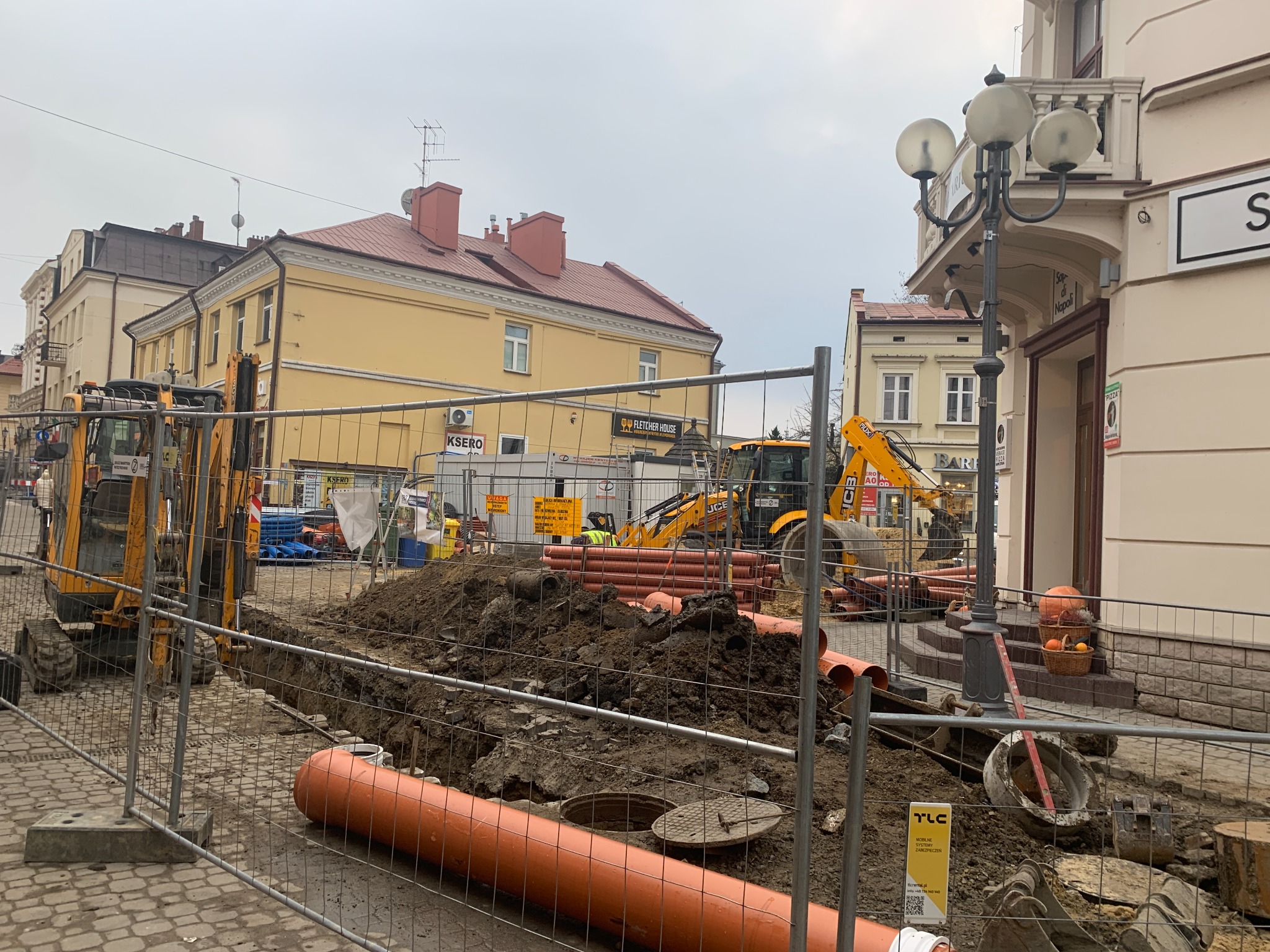 Żółte koparki przy wykopie z pomarańczowymi rurami na tle miejskiej zabudowy, zabezpieczone metalową siatką.