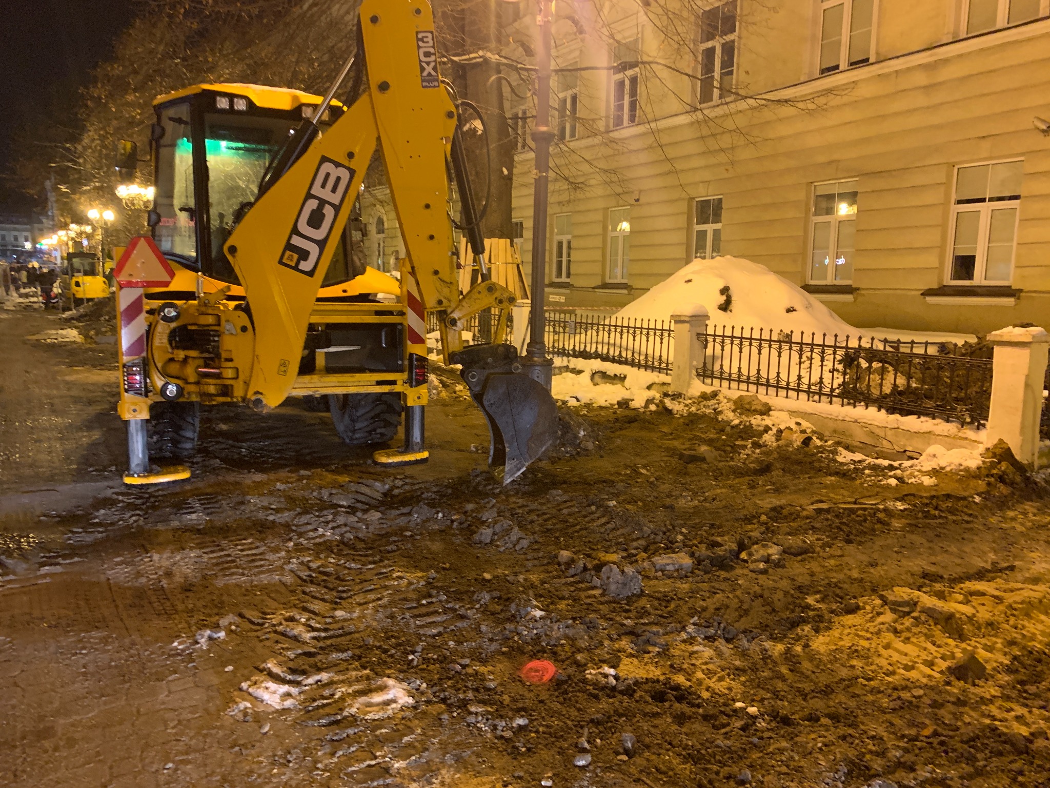 Żółta koparko-ładowarka JCB na błotnistej drodze z widocznymi śladami opon, w tle ośnieżony chodnik i budynek oświetlony latarniami ulicznymi nocą.