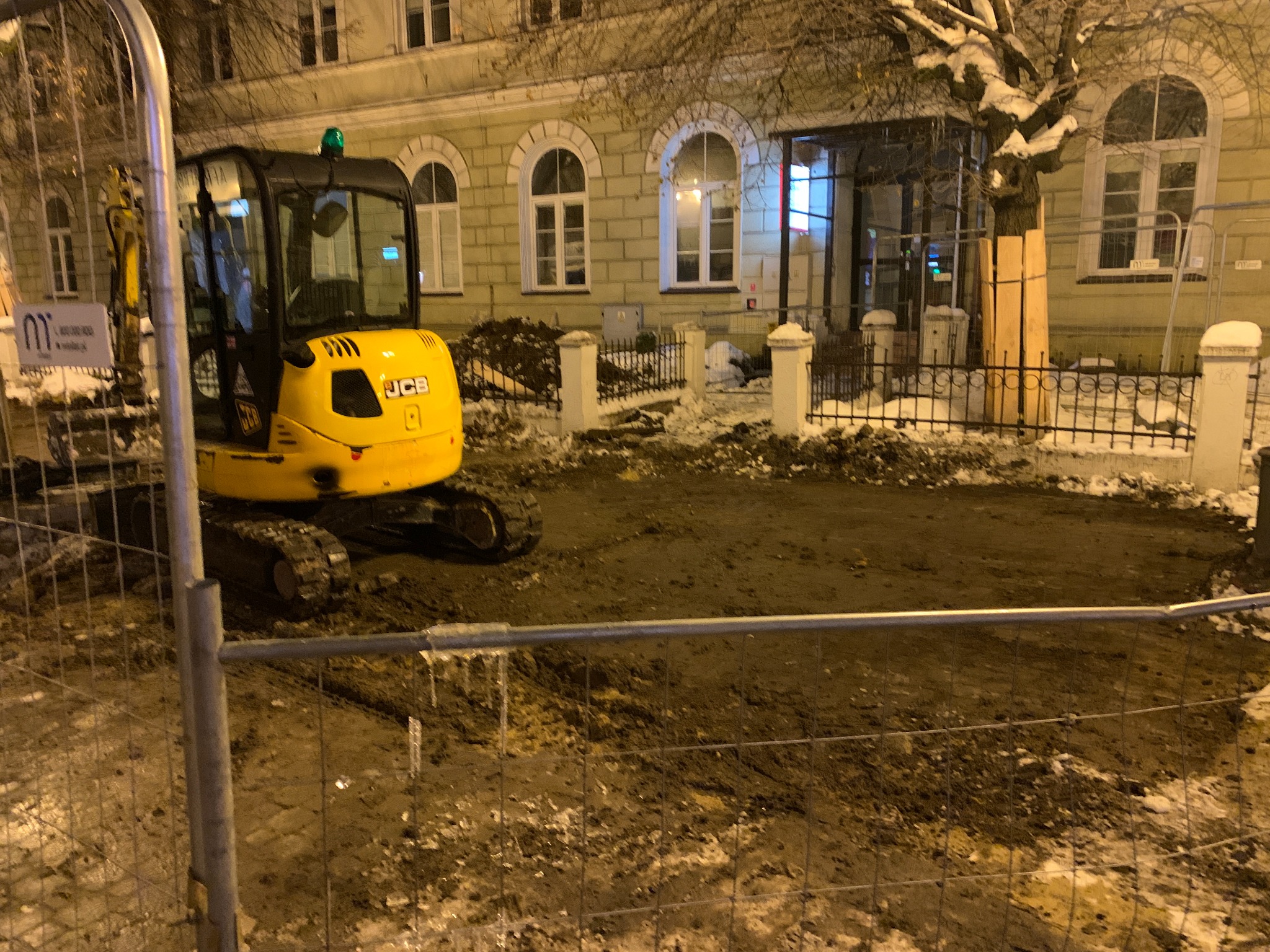 Żółta mini koparka JCB na placu budowy w zimowej scenerii, widoczna tymczasowa siatka zabezpieczająca, w tle budynek z oknami w kształcie łuków.