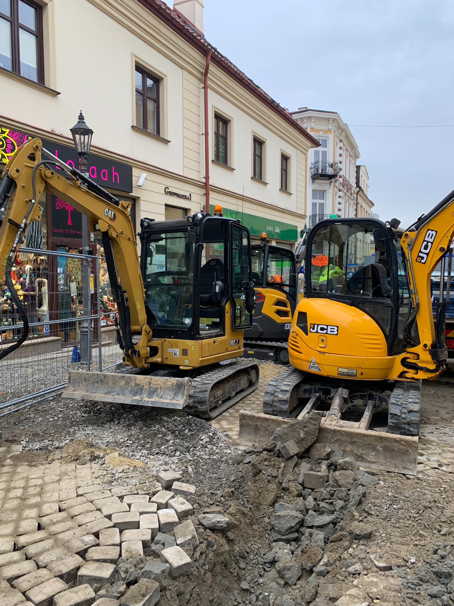 Dwa żółte minikoparki CAT i JCB na tle miejskiej ulicy z rozkopaną kostką brukową, widoczne budynki z szyldami sklepów.