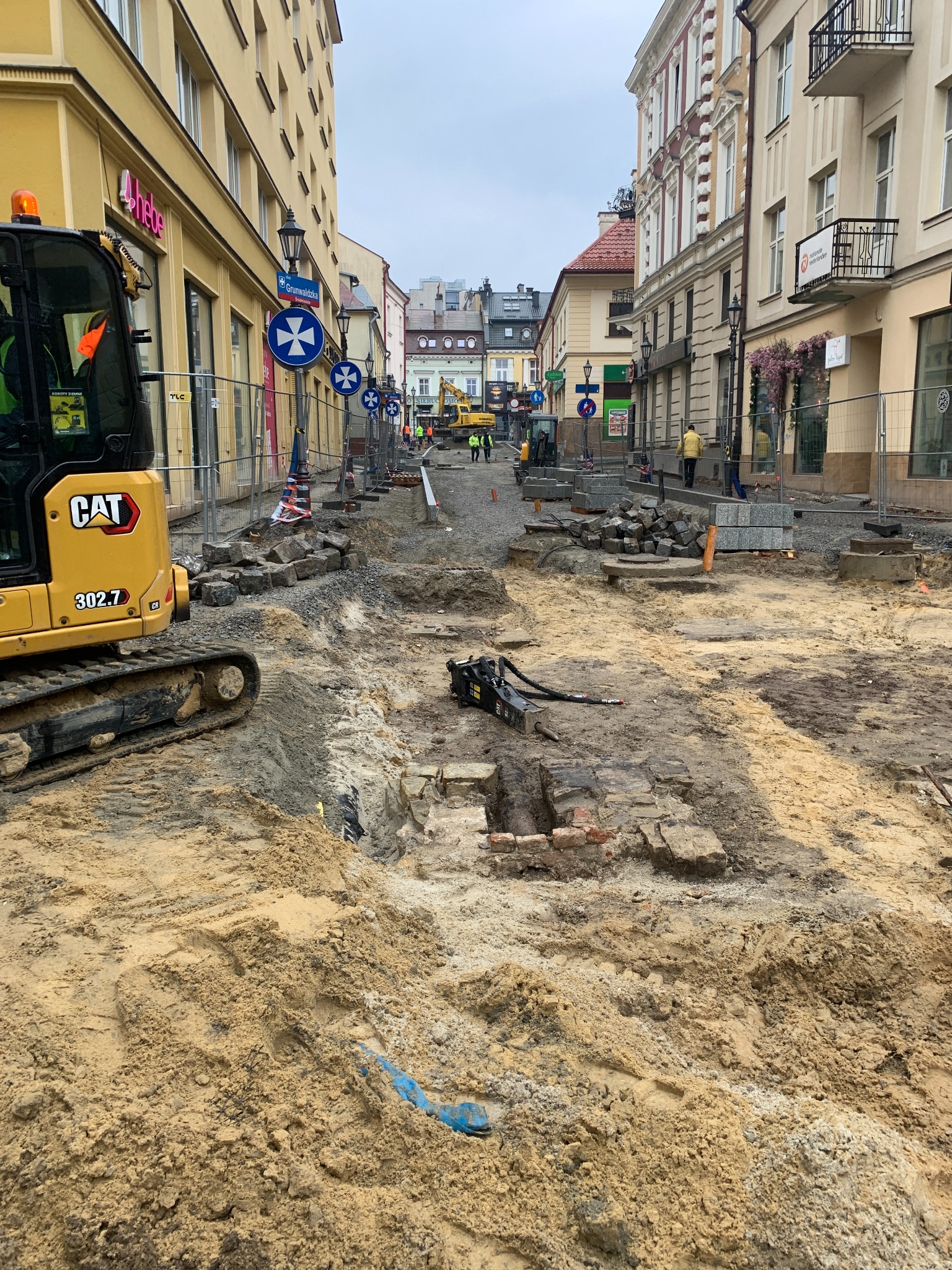 Ulica w trakcie remontu: rozkopana nawierzchnia, widoczny sprzęt budowlany CAT 302.7, robotnicy w oddali, znaki drogowe i budynki po obu stronach.
