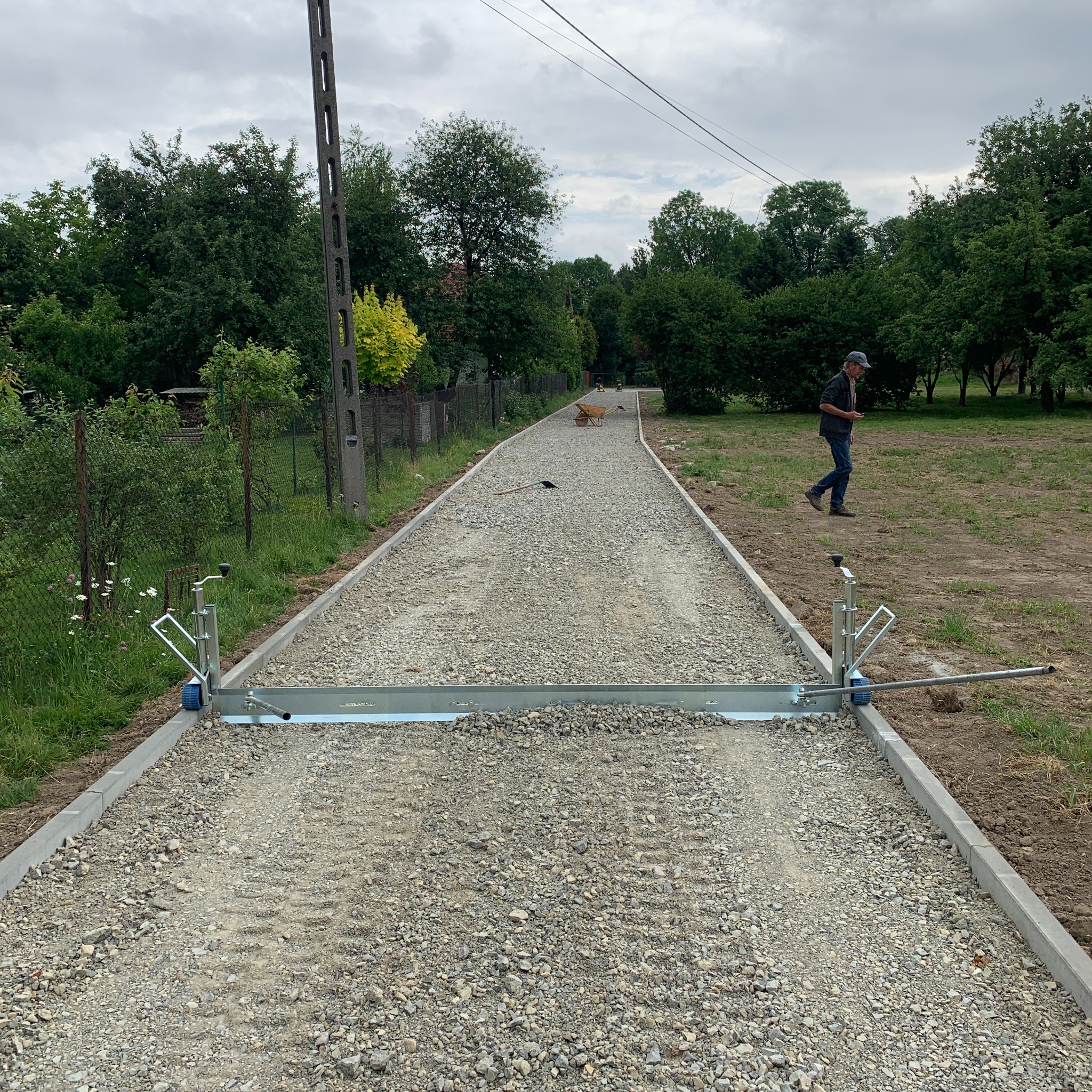 Świeżo utwardzona żwirowa droga z betonowymi obrzeżami, wyrównywana specjalistycznym narzędziem z rolkami, w tle widoczny mężczyzna i wózek budowlany.