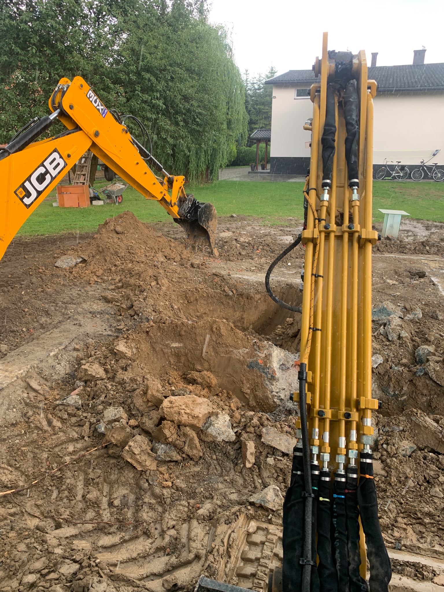 Żółta koparka JCB podczas prac ziemnych, widoczne ślady gąsienic, wykop, gruz i fragment budynku w tle.