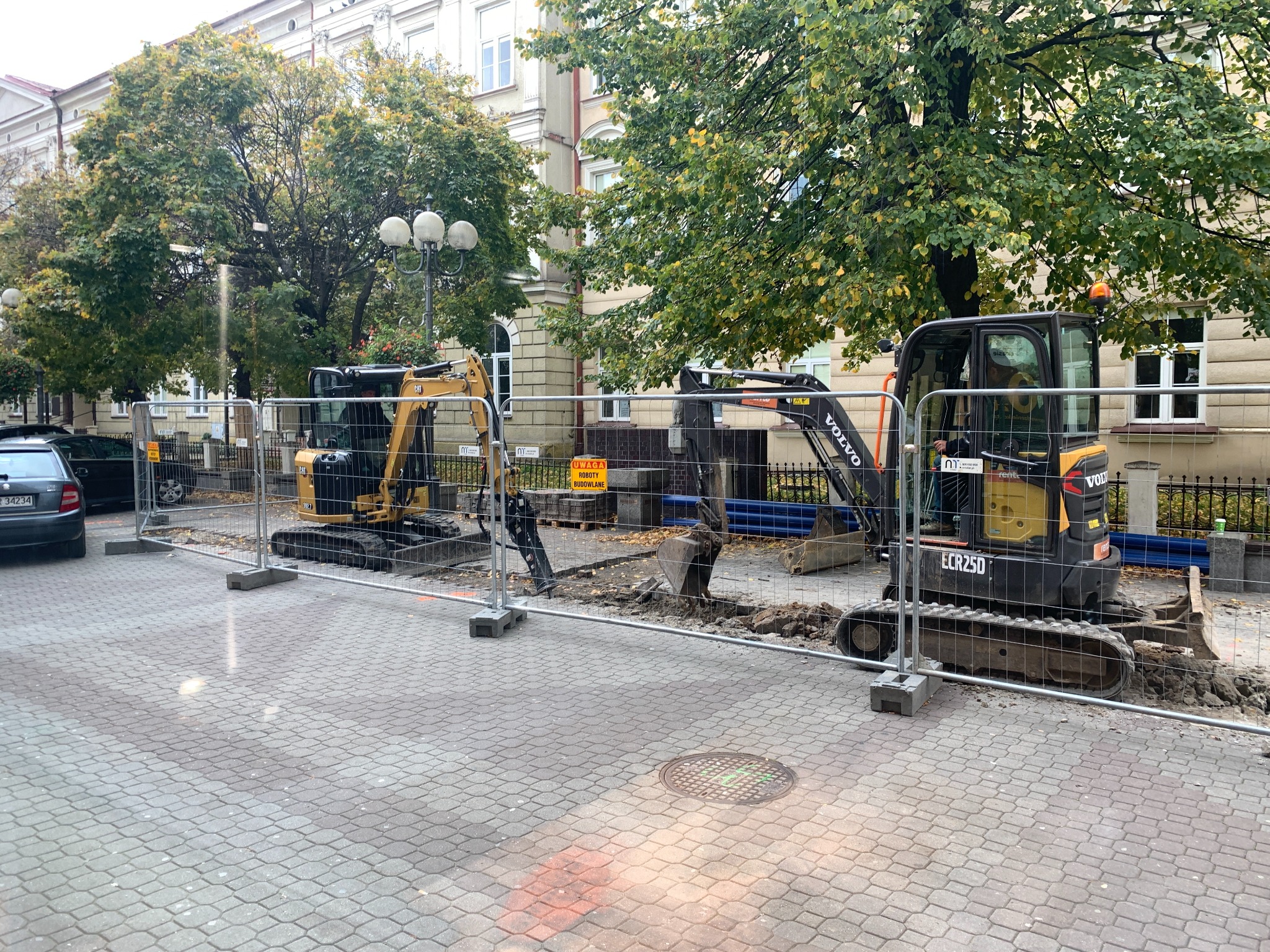 Dwa mini-koparki Volvo i CAT na kostce brukowej, zabezpieczone barierkami, prowadzone prace ziemne w otoczeniu drzew i budynków.