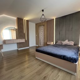 CLAVER MEBLE - Nowoczesna sypialnia z drewnianymi panelami ściennymi z podświetleniem LED, okrągłym lustrem z podświetleniem, łóżkiem z szarą narzutą i stolikami nocnymi w kolorze pudrowego różu.