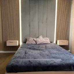 Nowoczesna sypialnia z łóżkiem w kolorze pudrowego różu, szarą tapicerowaną ścianą z podświetleniem LED i drewnianymi panelami lamelowymi po bokach, minimalistyczna lampa wisząca.