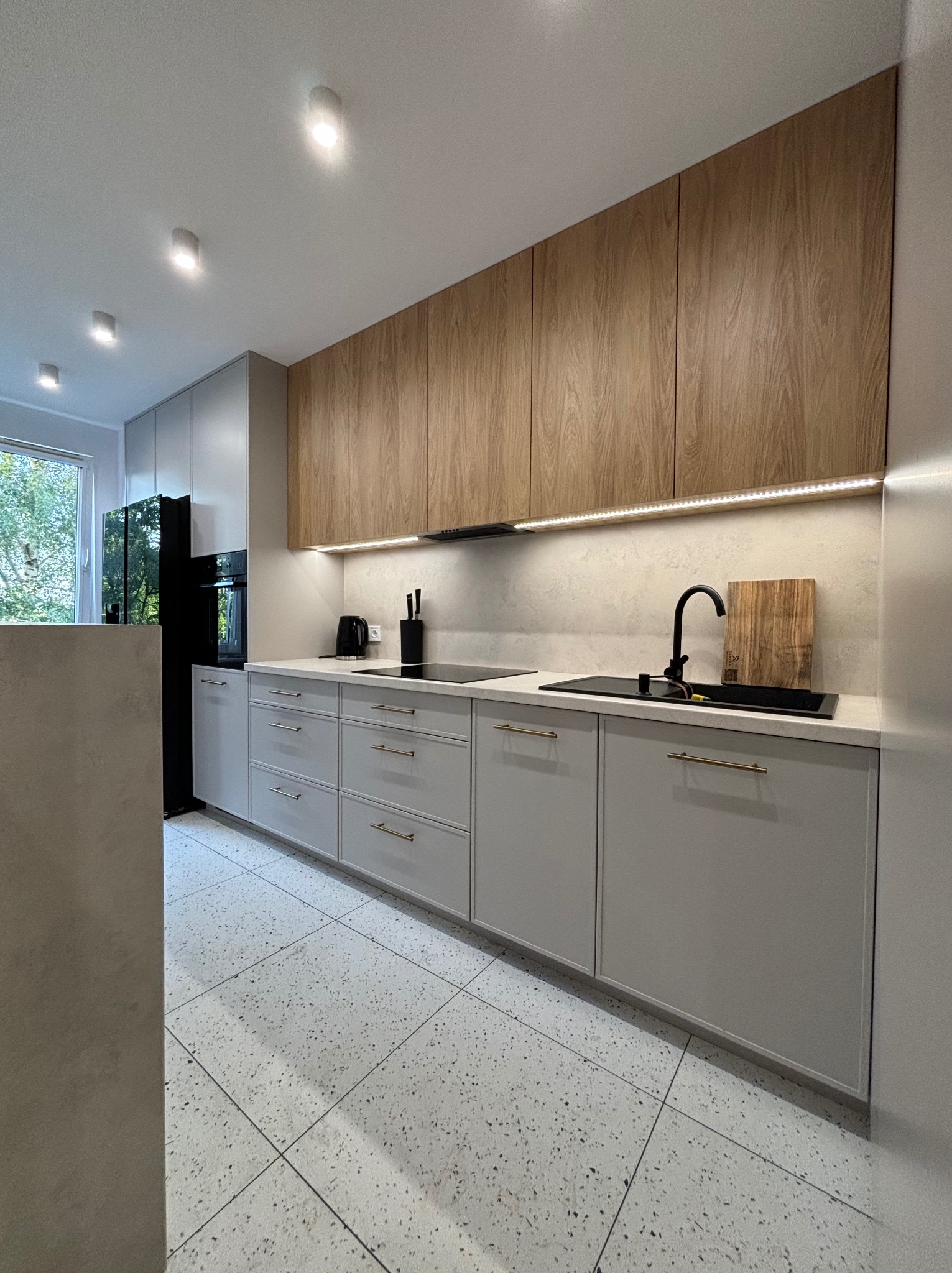 Nowoczesna kuchnia: szare szafki dolne z poziomymi, złotymi uchwytami, drewniane szafki górne z oświetleniem LED podszafkowym, czarny zlew i bateria, podłoga w stylu lastryko.