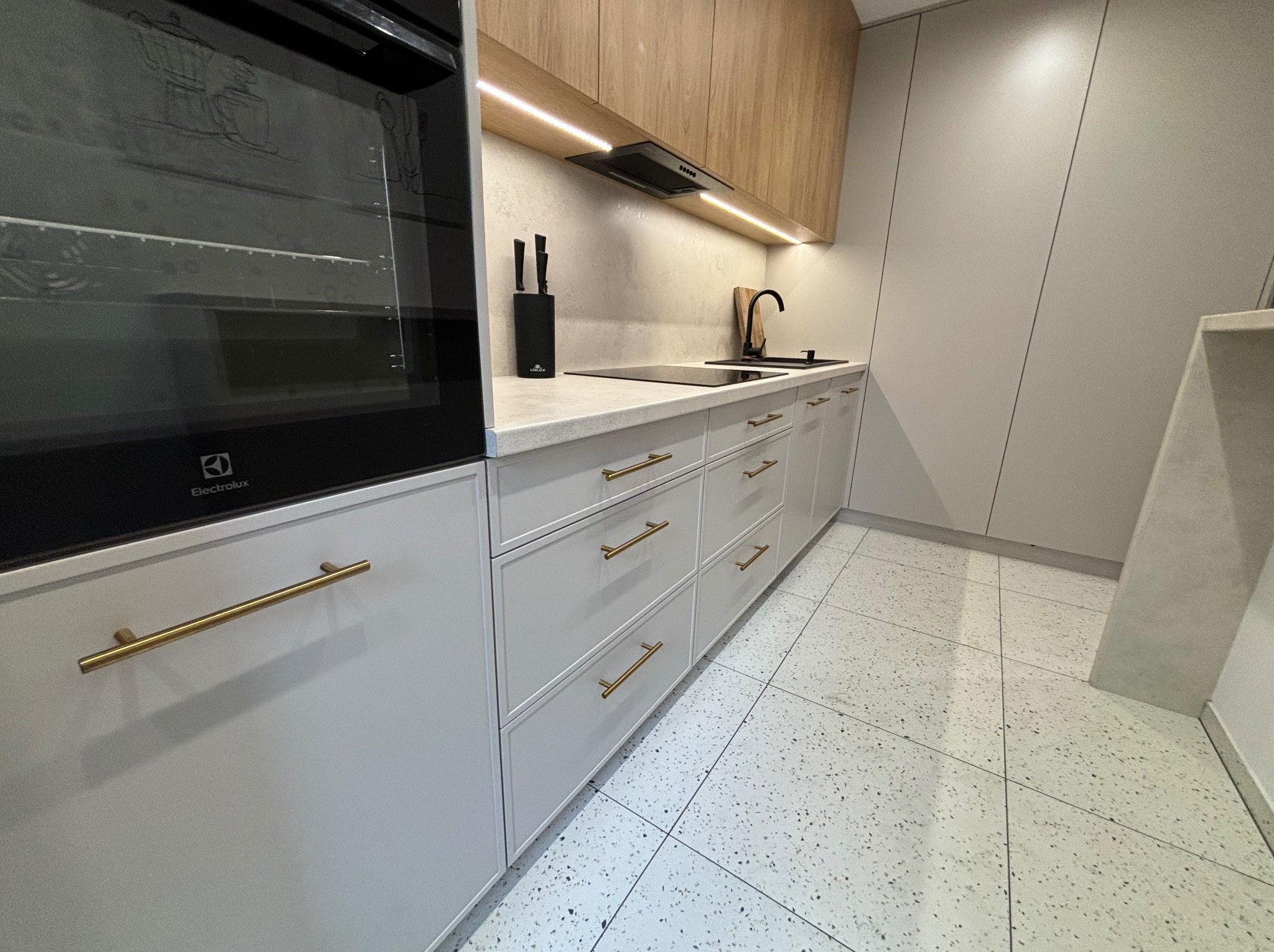 Nowoczesna kuchnia z frontami w odcieniach szarości i beżu, złotymi uchwytami, płytą indukcyjną, czarną baterią i podświetleniem LED pod szafkami wiszącymi, widoczny piekarnik Electrolux po lewej...