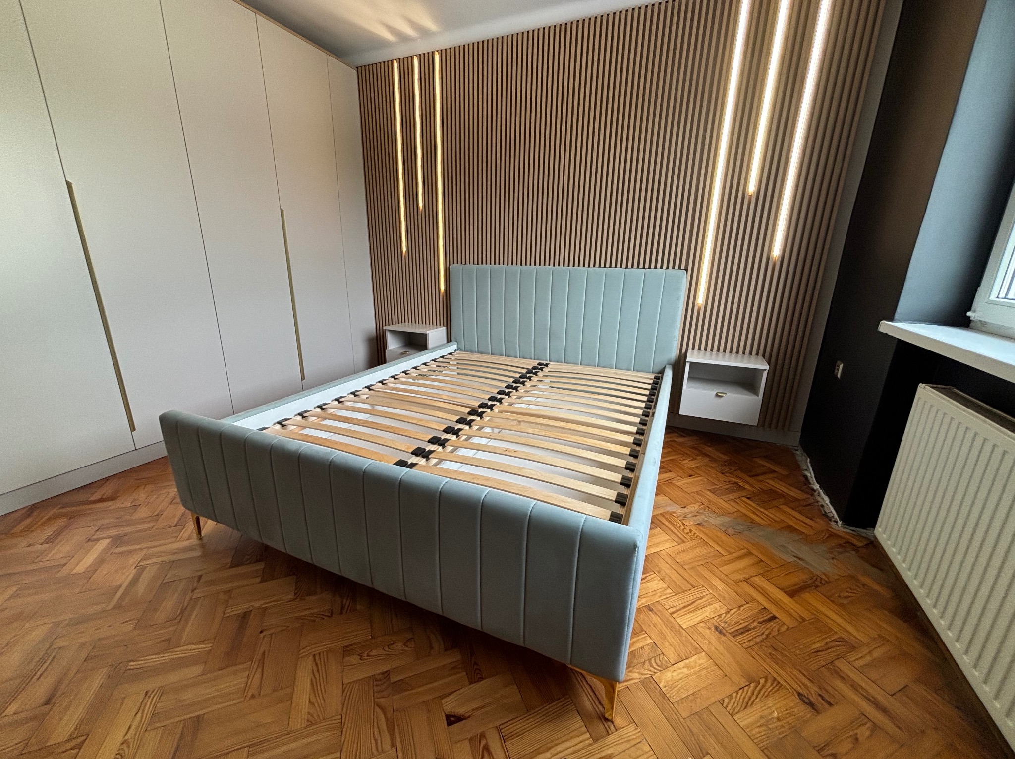 Łóżko z tapicerowanym zagłówkiem w odcieniach szarości, ustawione na parkiecie w jodełkę, z szafą w tle i dekoracyjną ścianą z drewnianych paneli z oświetleniem LED.