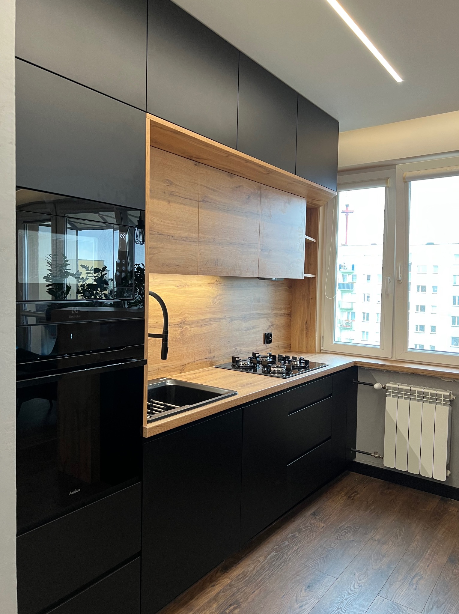 Nowoczesna kuchnia: czarne matowe szafki kontrastują z drewnianym blatem i panelami, widoczny piekarnik Amica, płyta gazowa i czarny zlew, a w oknie widok na blok mieszkalny.