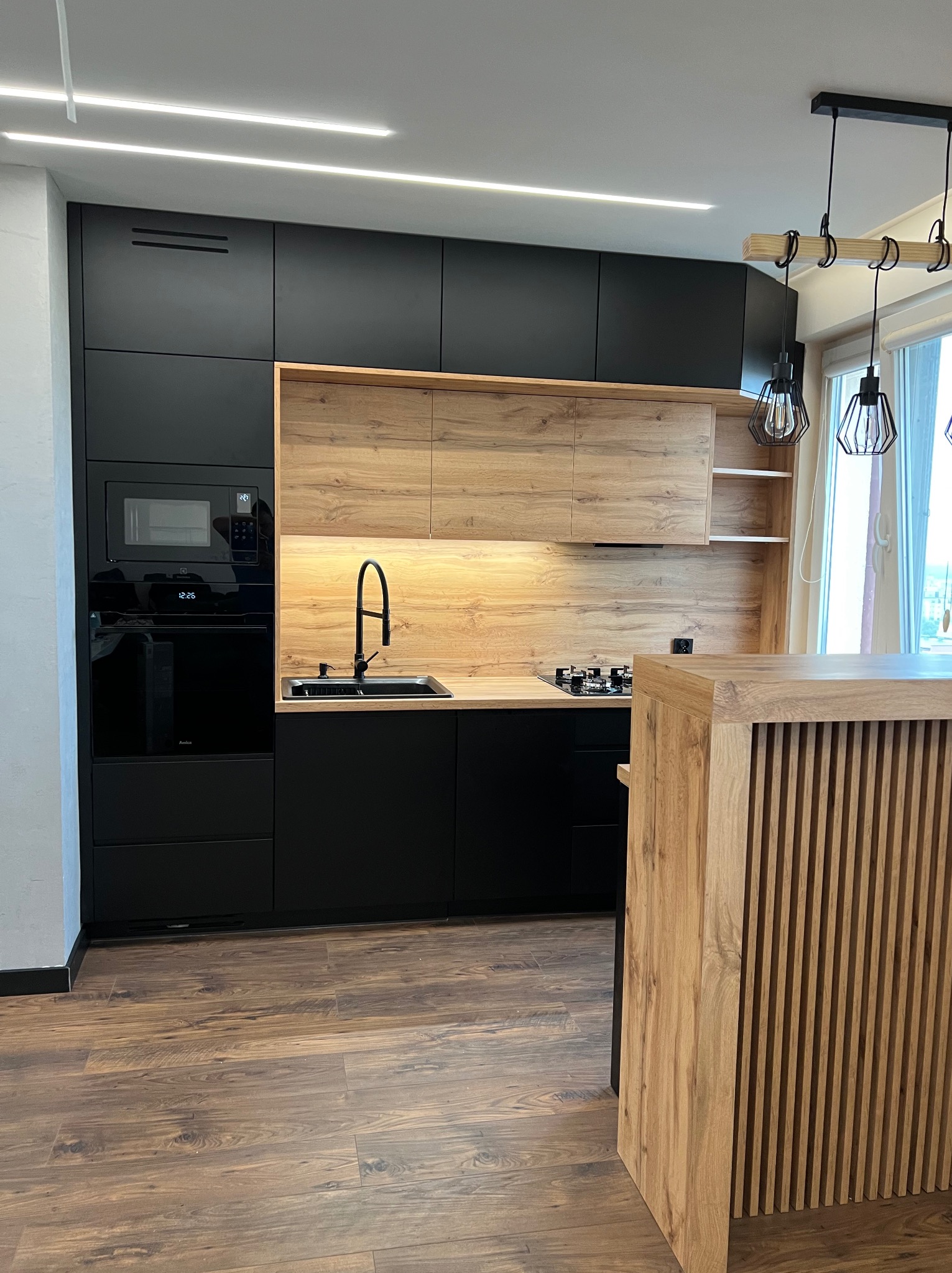 Nowoczesna kuchnia: czarne matowe szafki dolne i górne, drewnopodobne szafki wiszące z podświetleniem LED, czarny zlew i bateria, zabudowany piekarnik i mikrofalówka, drewniana wyspa z pionowymi...
