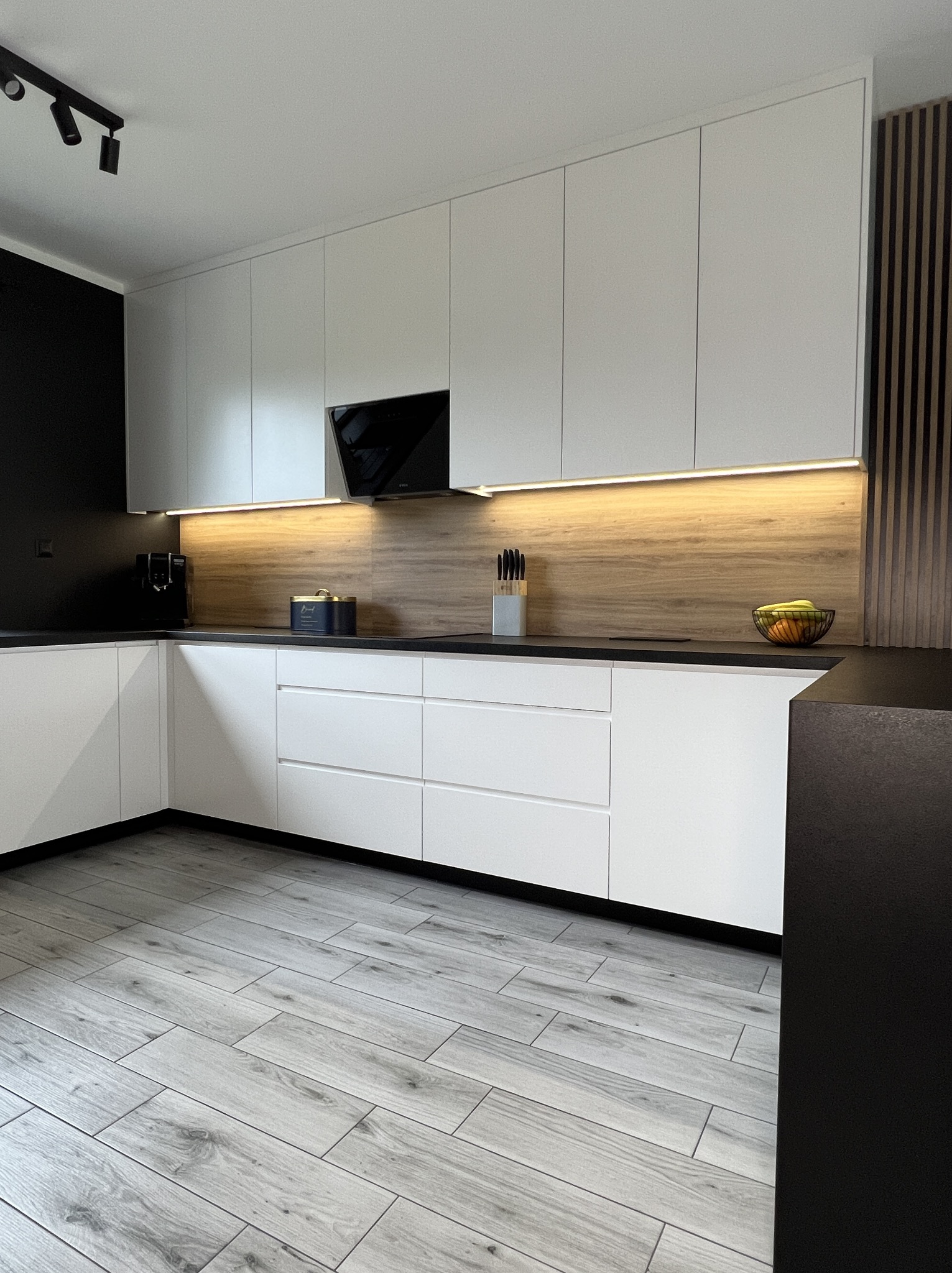 Nowoczesna kuchnia z białymi, gładkimi frontami szafek, czarnym blatem i drewnopodobnym panelem ściennym z oświetleniem LED, widok na drewniane panele dekoracyjne i jasnoszarą podłogę imitującą...