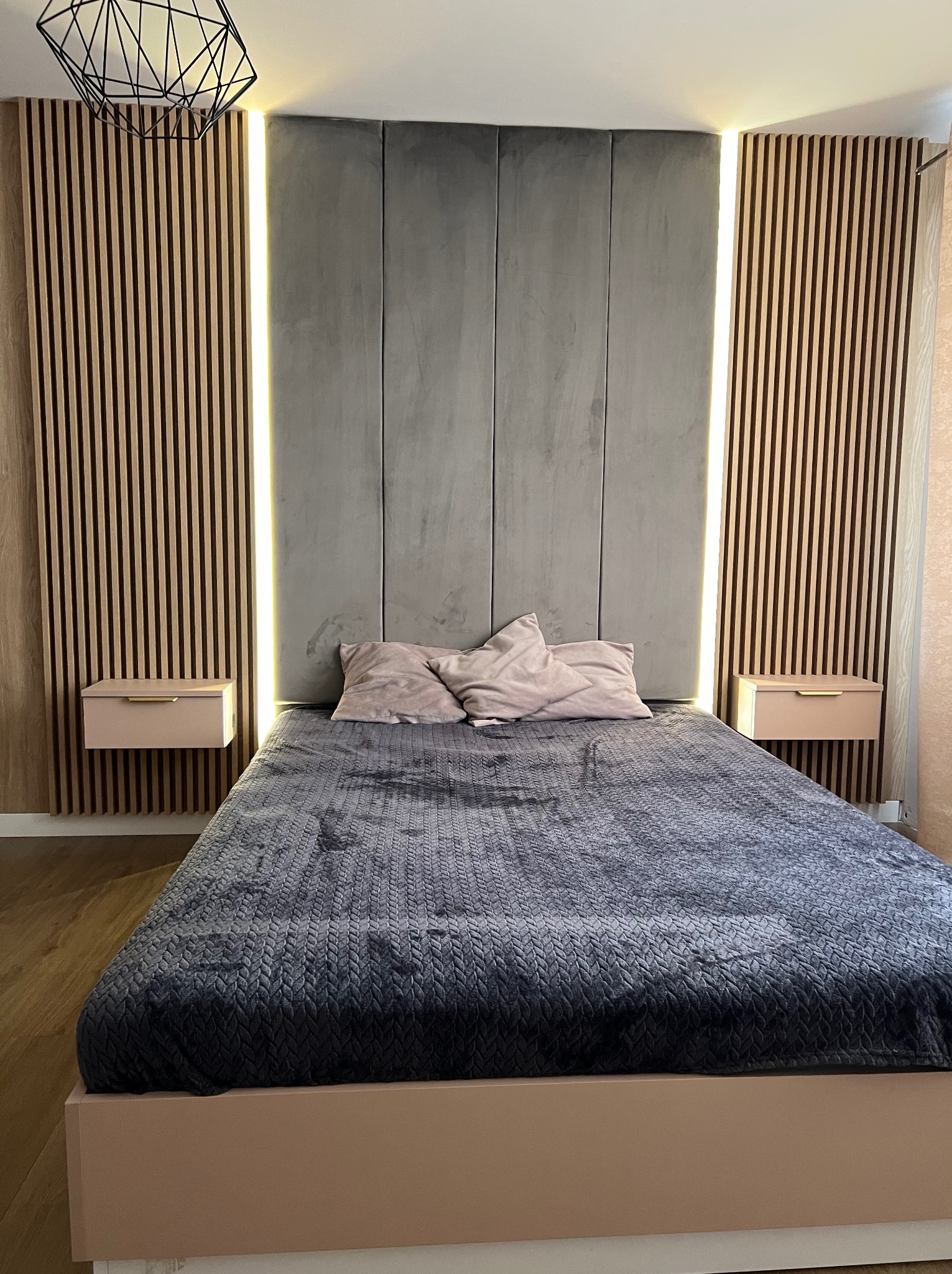 Nowoczesna sypialnia z łóżkiem w kolorze pudrowego różu, szarą tapicerowaną ścianą z podświetleniem LED i drewnianymi panelami lamelowymi po bokach, minimalistyczna lampa wisząca.