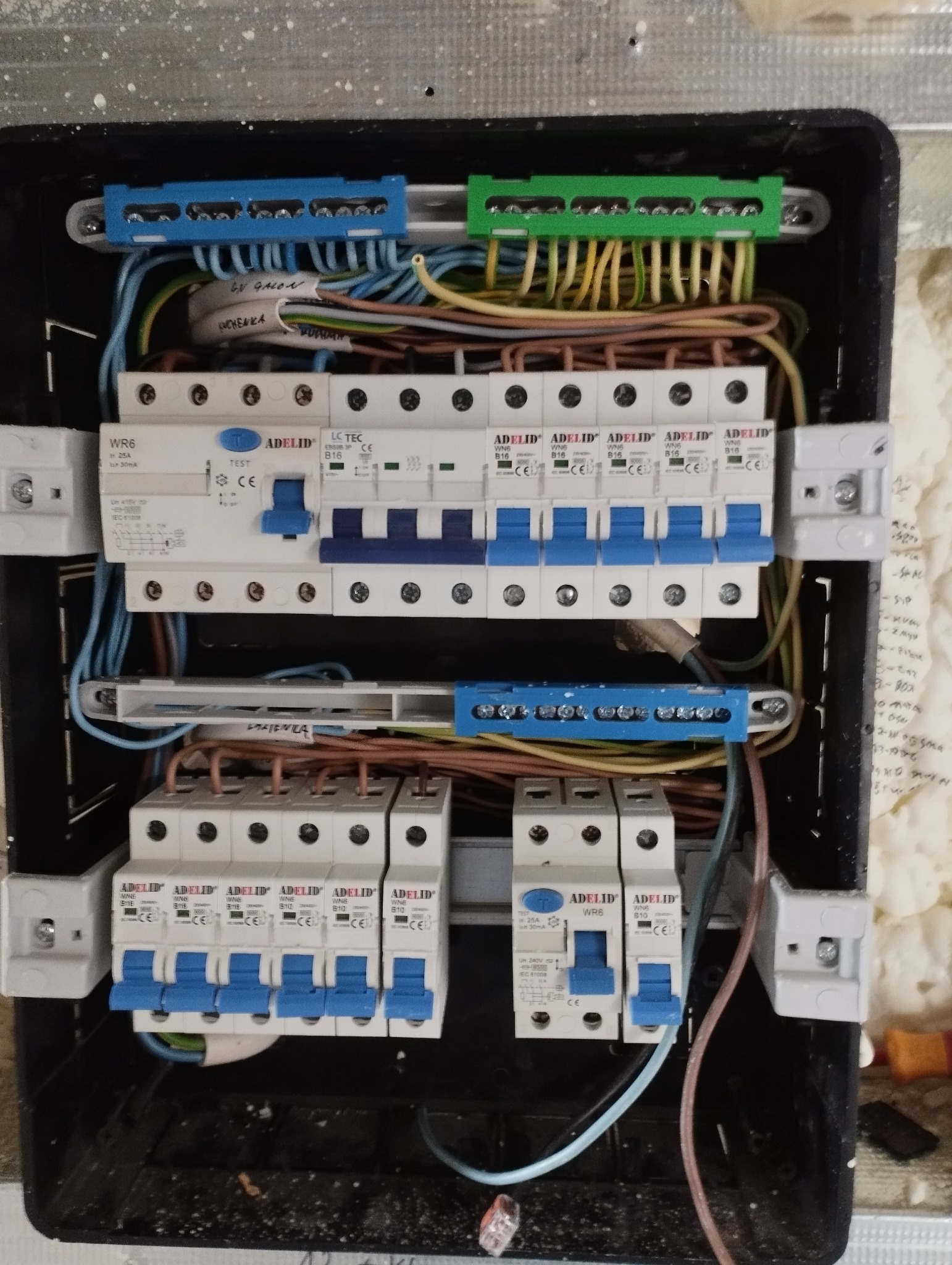 Tablica rozdzielcza z bezpiecznikami i okablowaniem zamontowana w ścianie w Poznaniu, widoczne listwy zaciskowe w kolorach niebieskim i zielonym oraz opisane przewody.