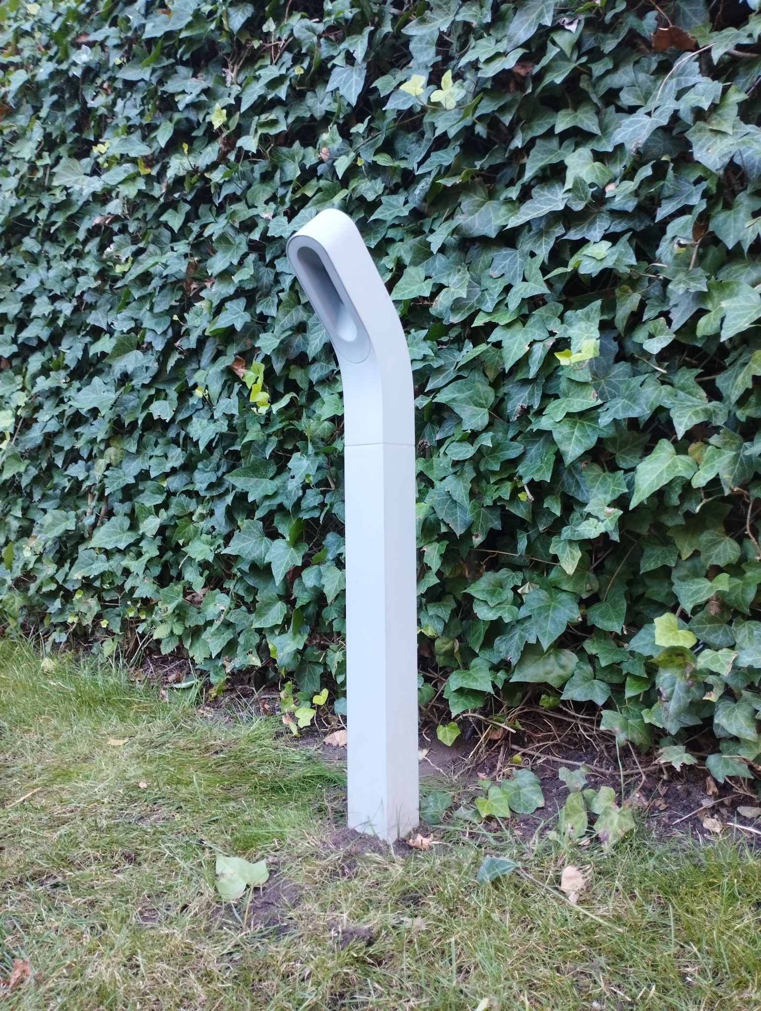 Nowoczesna, szara lampa ogrodowa typu słupek oświetlająca trawnik przed ścianą zielonego bluszczu.