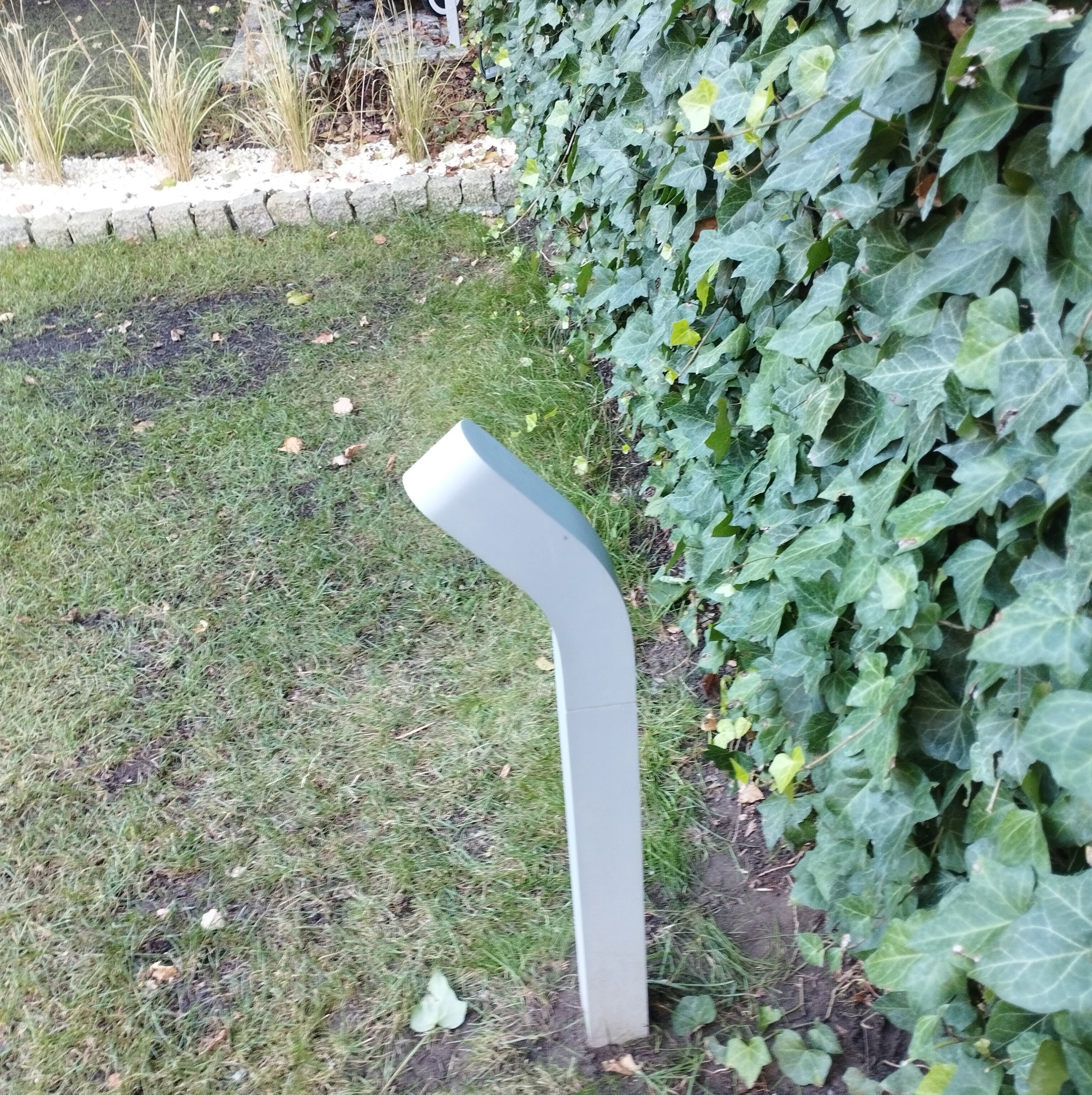 Nowoczesna lampa ogrodowa w kształcie litery L, wykonana z szarego tworzywa, oświetlająca trawnik i ścianę z bluszczu.