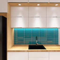 Nowoczesna kuchnia z białymi szafkami, drewnianym blatem i turkusowymi płytkami, czarny zlew i lodówka, oświetlenie podszafkowe LED.