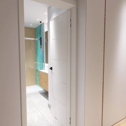 Art Tabor Design Marta Tabor - Widok na łazienkę z kabiną prysznicową z elementami dekoracyjnymi w odcieniach turkusu, umywalką na drewnianej szafce i białymi płytkami podłogowymi, widzianą z korytarza z białą zabudową szaf.