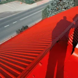 Czerwony dach z blachy trapezowej, cień dwóch osób, widok z góry na ulicę i drzewa.