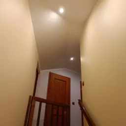 Klatka schodowa z drewnianą barierką i poręczą, ściany pomalowane na jasny beż, widoczne drzwi na p&oacute;łpiętrze i oświetlenie sufitowe.