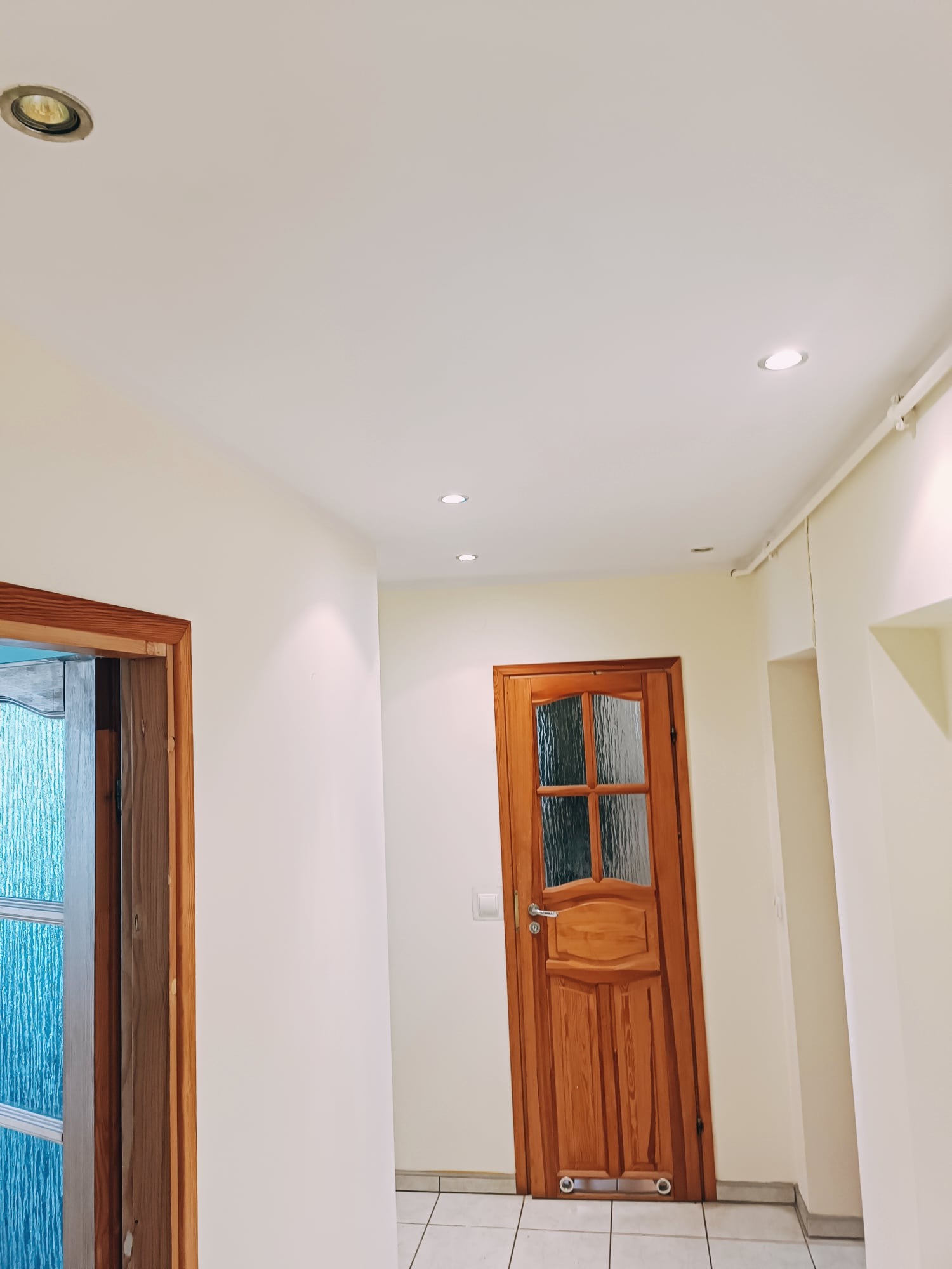 Wnętrze mieszkania z jasnymi ścianami, drewnianą stolarką drzwiową i punktowym oświetleniem sufitowym. Widoczny fragment korytarza z dwoma parami drzwi.