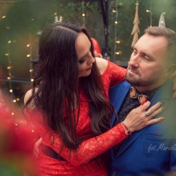 JORDAN MARIOLA - Elegancka para: kobieta w czerwonej koronkowej sukience obejmuje mężczyznę w niebieskiej marynarce i drewnianej muszce, oboje patrzą na siebie w romantycznej atmosferze z dekoracyjnymi światełkami...