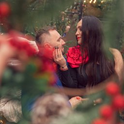 JORDAN MARIOLA - Zakochana para w eleganckich strojach, kobieta w czarnej sukni z czerwoną ozdobą, mężczyzna w granatowej marynarce, patrząca na siebie w scenerii świątecznej, widok przez gałązki z czerwonymi owocami.