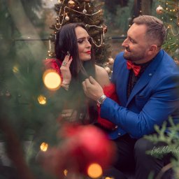 JORDAN MARIOLA - Elegancka para siedzi na tle choinki, kobieta w czerwonej sukni i mężczyzna w niebieskiej marynarce z czerwoną muszką, patrzą na siebie z uśmiechem w świątecznej atmosferze.