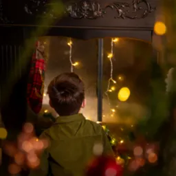 Chłopiec w zielonej koszuli siedzi tyłem, patrząc na kominek udekorowany świątecznymi lampkami i ozdobami, w tle widać dekoracje świąteczne na kominku.