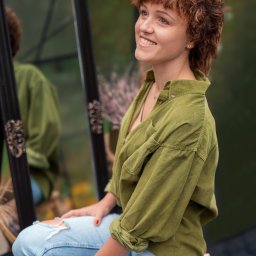 JORDAN MARIOLA - Uśmiechnięta kobieta z rudymi kręconymi włosami, w zielonej koszuli i podartych jeansach, siedzi na stołku obok lustra, z zamyślonym spojrzeniem skierowanym w górę.