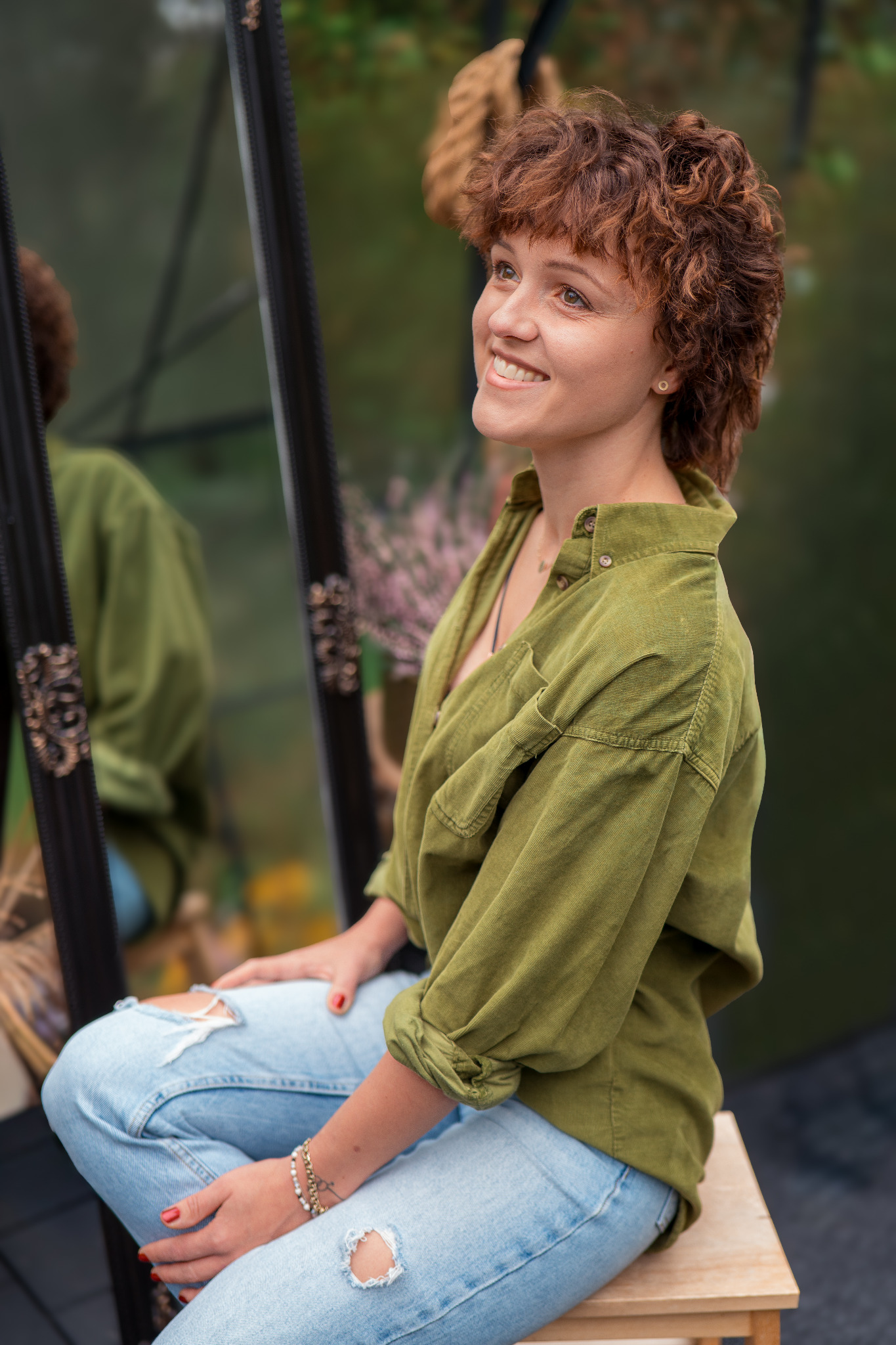 Uśmiechnięta kobieta z rudymi kręconymi włosami, w zielonej koszuli i podartych jeansach, siedzi na stołku obok lustra, z zamyślonym spojrzeniem skierowanym w górę.