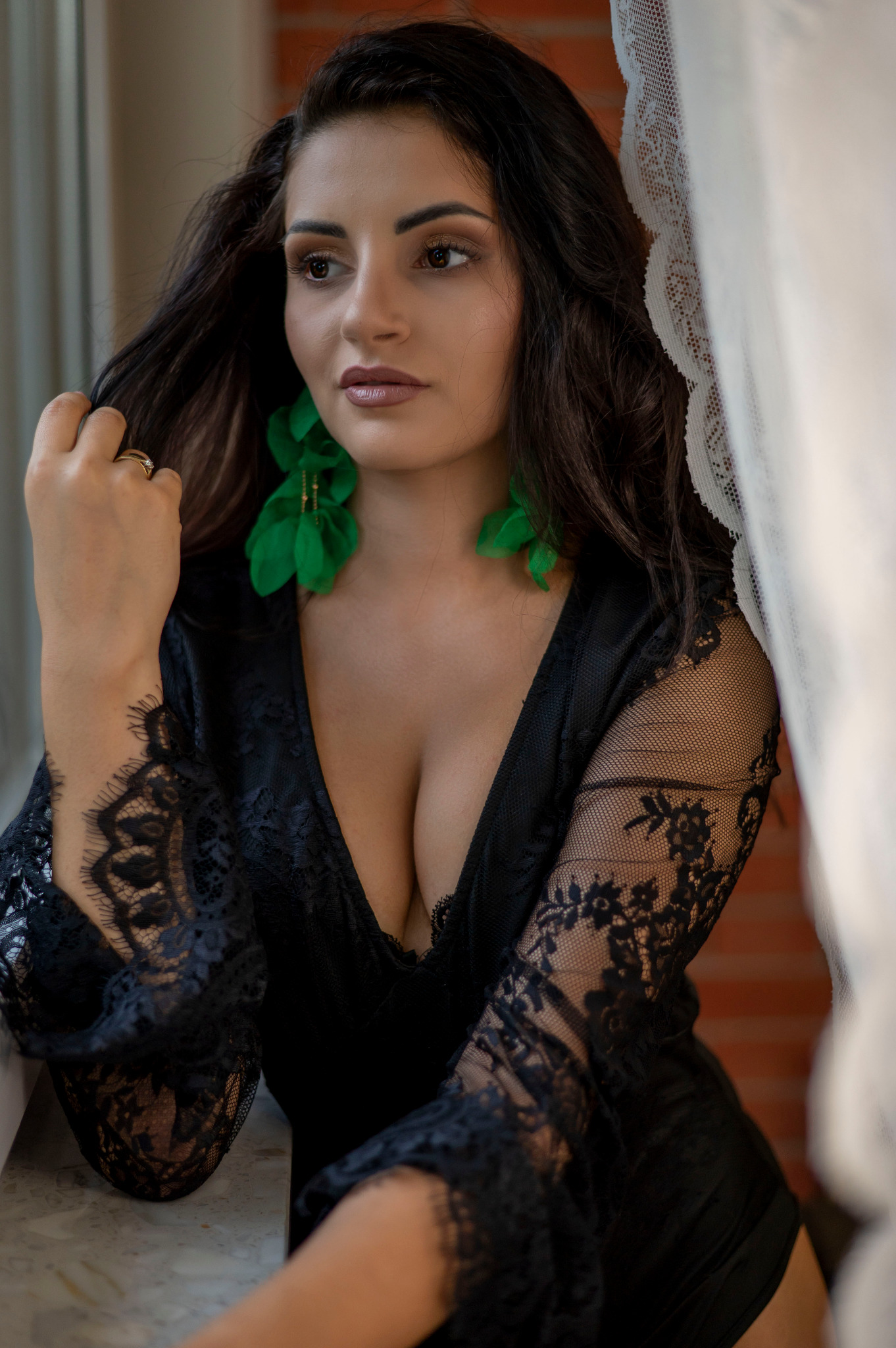 Kobieta w czarnej koronkowej bieliźnie, z zielonymi kolczykami, siedzi przy oknie z firanką, spoglądając w bok. Delikatne światło podkreśla jej urodę.