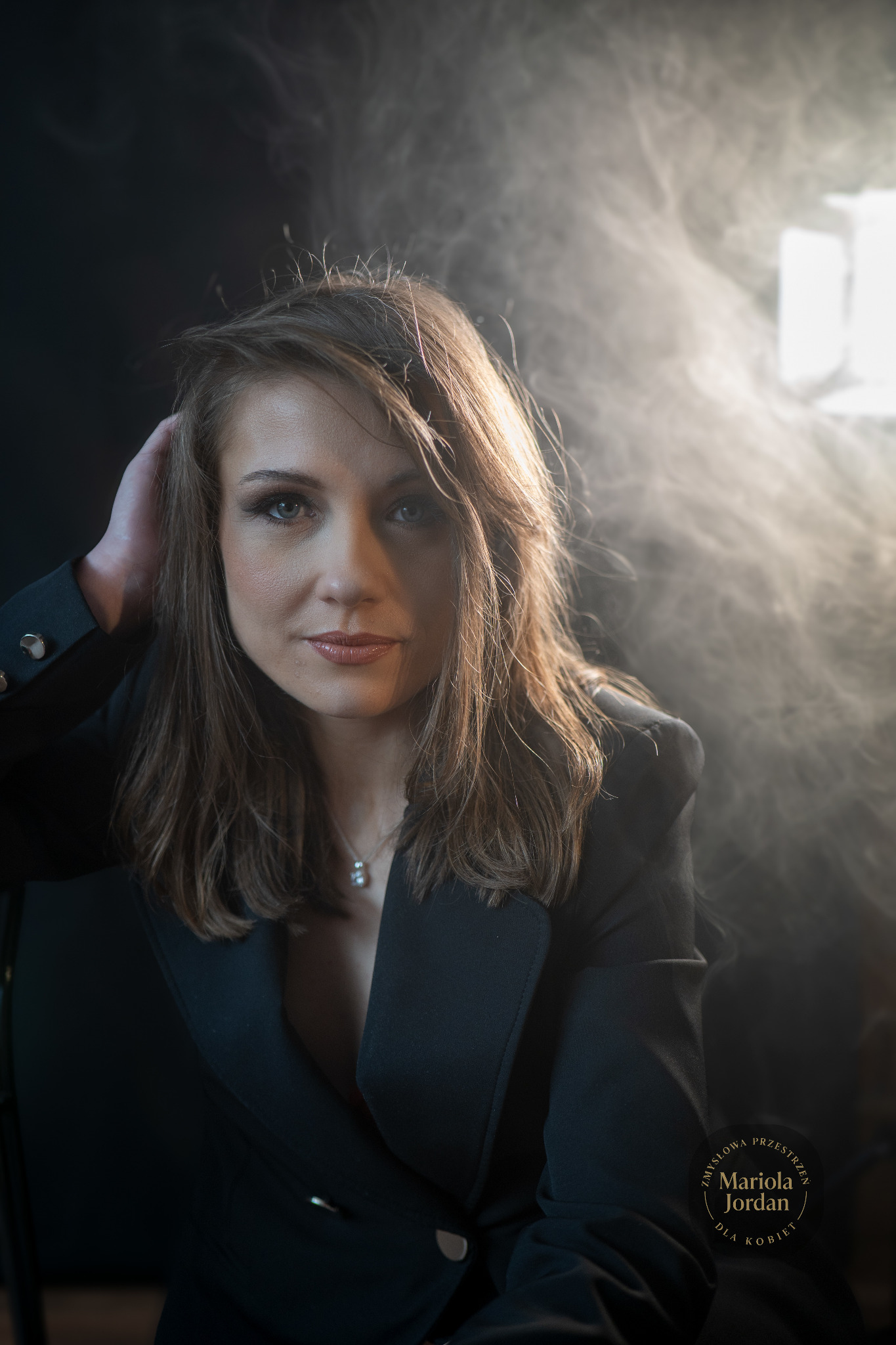 Portret kobiety w czarnej marynarce na ciemnym tle z efektem dymu i oświetleniem studyjnym, uchwycony w półzbliżeniu z ręką we włosach i delikatnym uśmiechem.