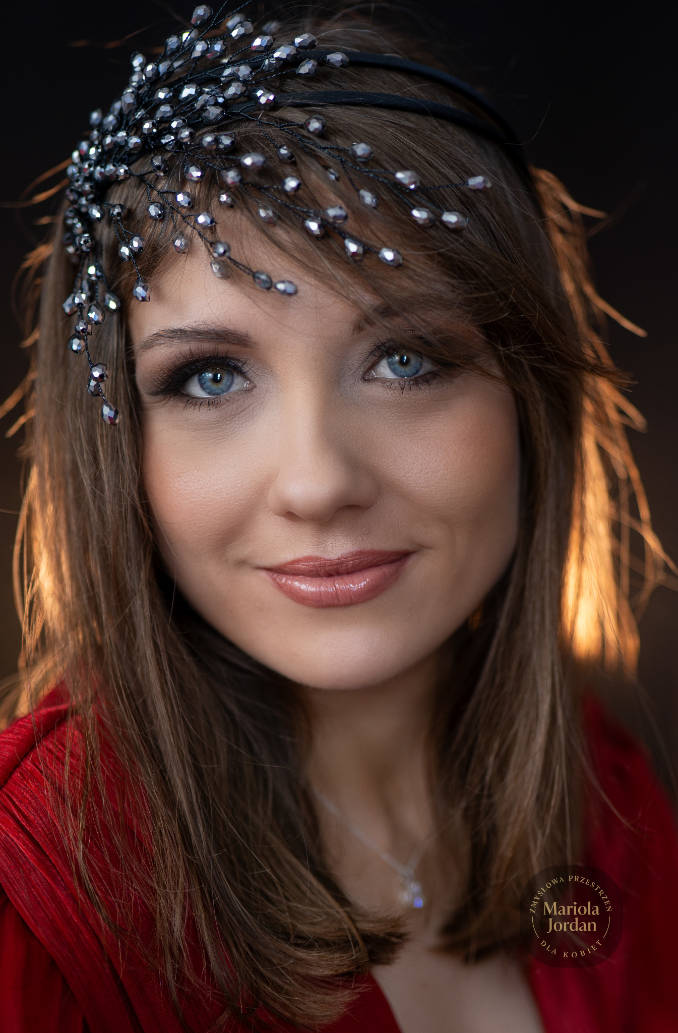 Portret kobiety z niebieskimi oczami, w czerwonej sukience i ozdobą na głowie z koralików, uśmiechająca się do kamery na ciemnym tle.