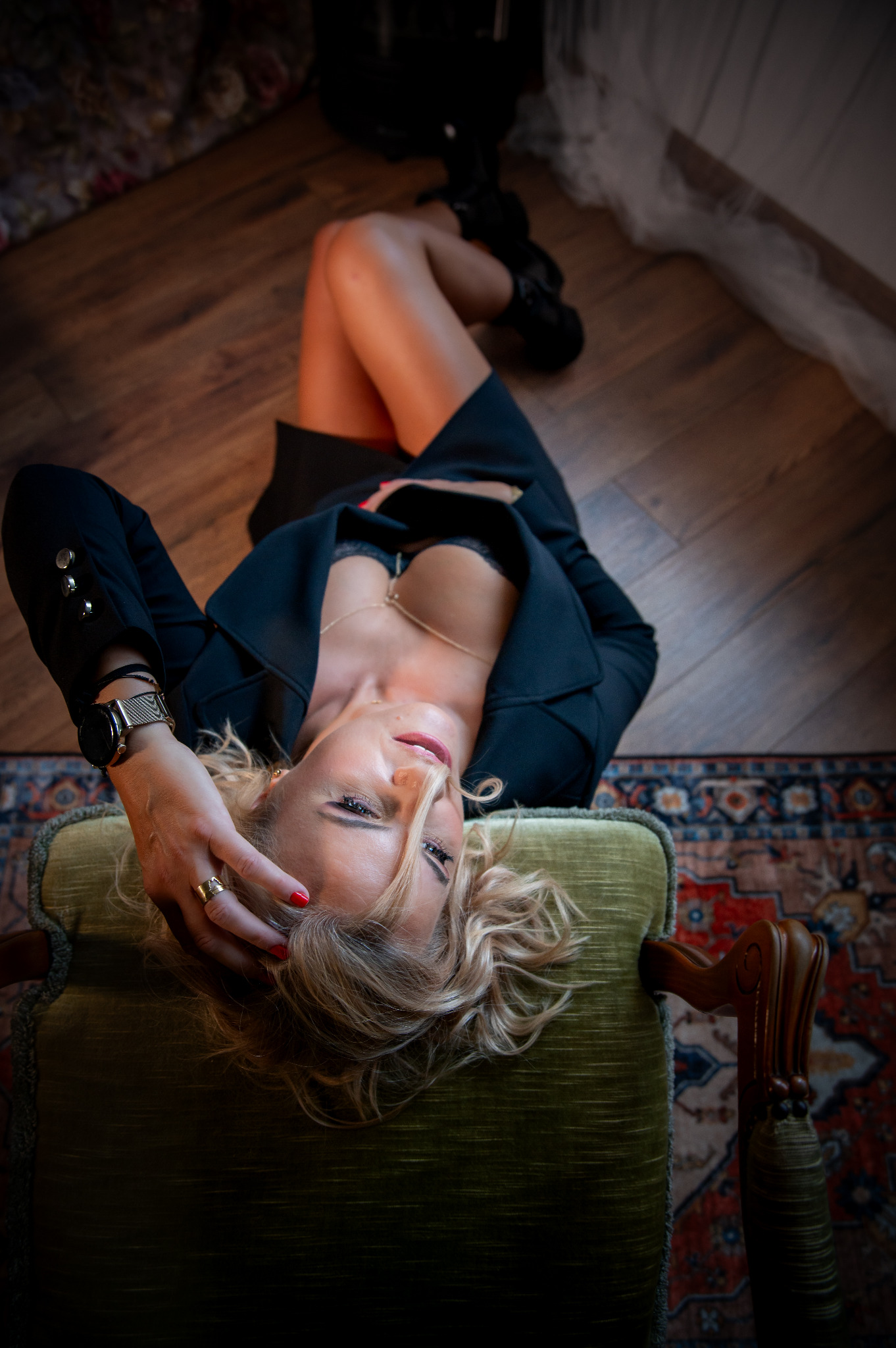 Kobieta w czarnej marynarce i bieliźnie leży na zielonym fotelu, patrząc w górę. Widoczny zegarek na ręce i pierścionek. Ujęcie z góry, na drewnianej podłodze.