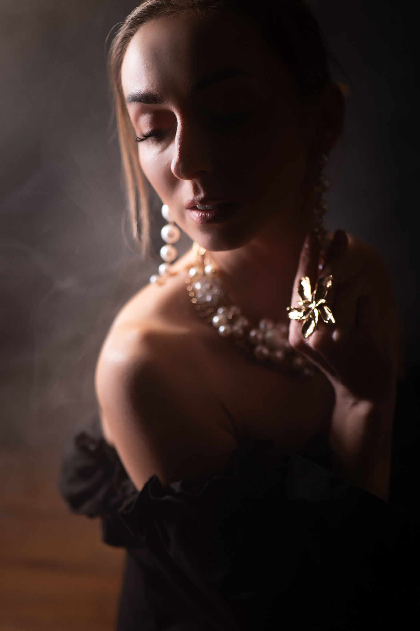 Portret kobiety w ciemnym oświetleniu, z perłami i złotym pierścieniem w kształcie kwiatu. Subtelny makijaż i elegancka stylizacja.