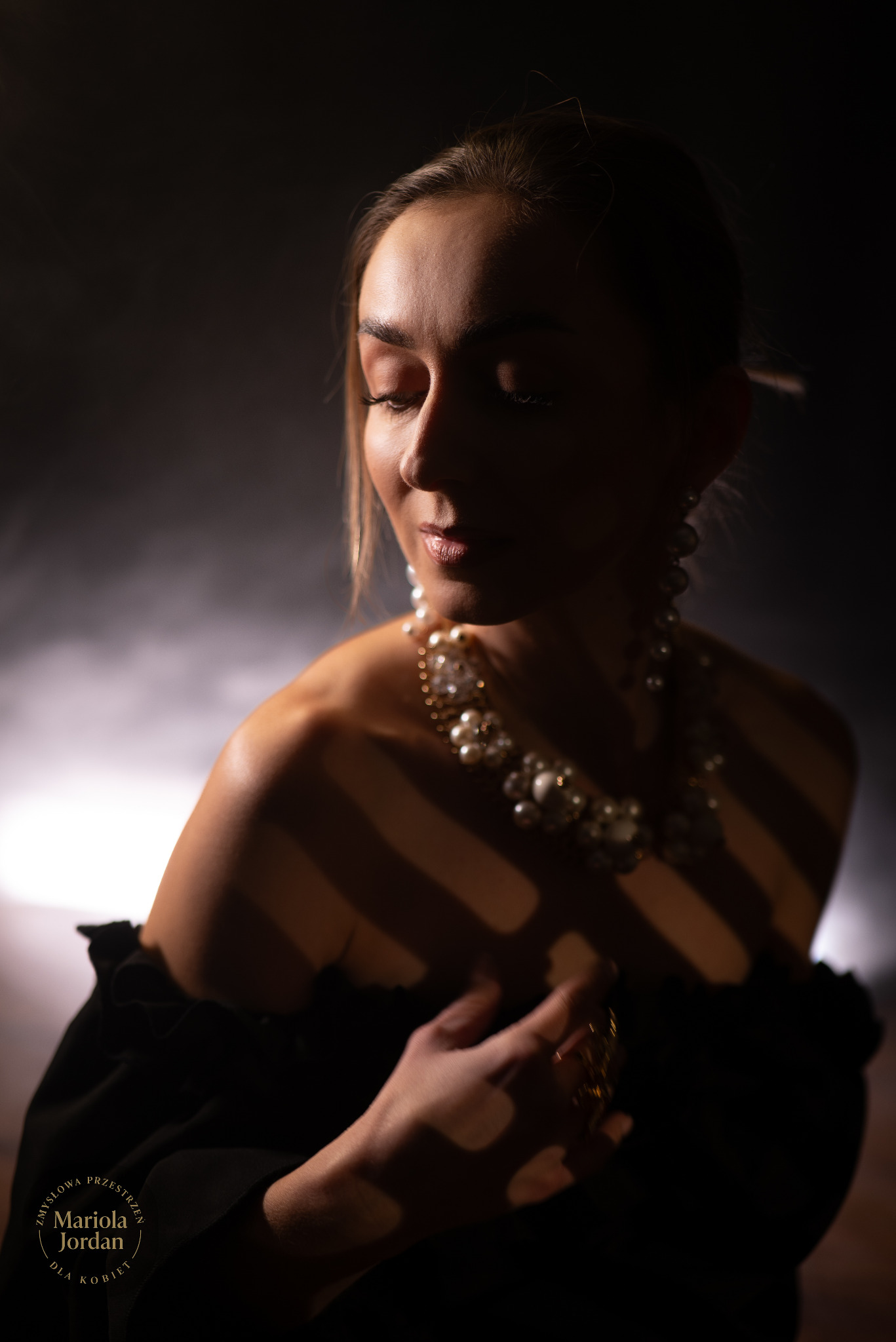 Portret kobiety w czarnej sukni z perłowym naszyjnikiem, oświetlonej paskami światła, na ciemnym tle. Zamknięte oczy, subtelny uśmiech, elegancki styl.