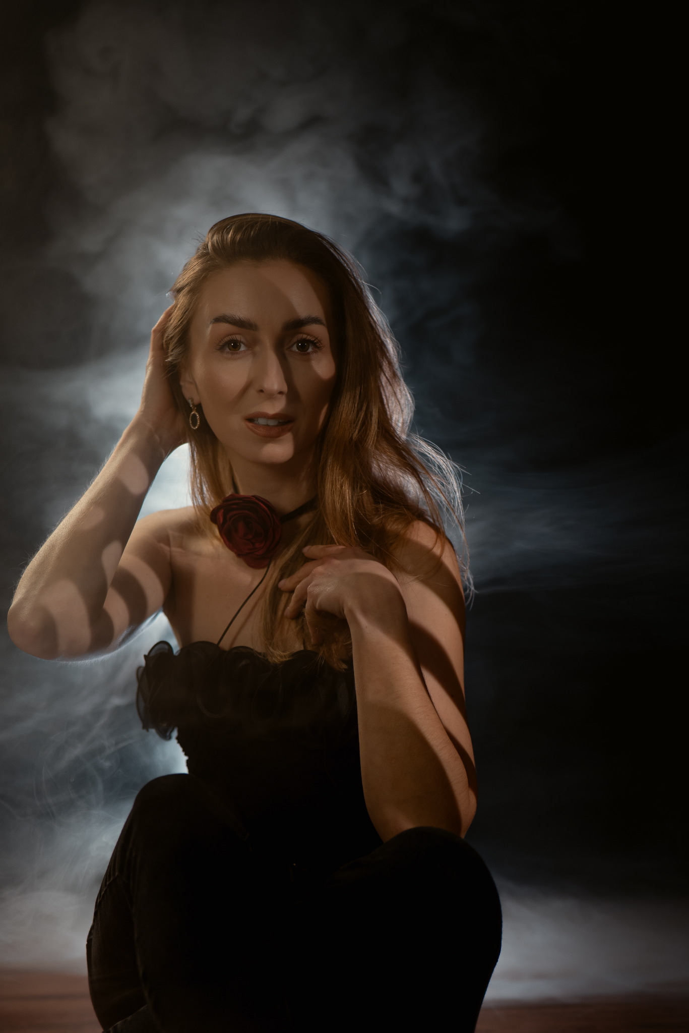 Portret kobiety w czarnej sukni z różą na szyi, pozującej w dymie i cieniach. Ujęcie studyjne z artystycznym oświetleniem.