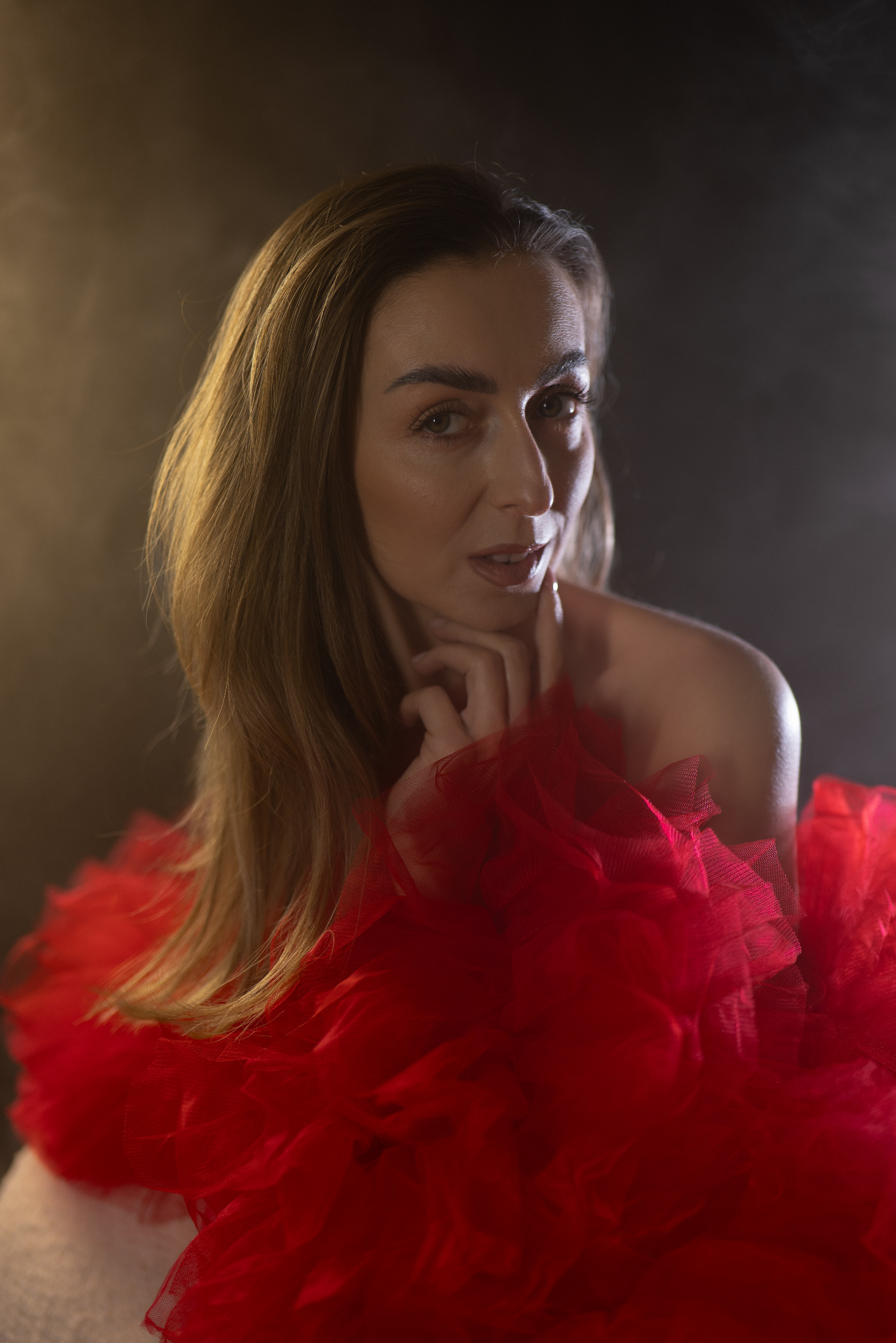 Portret kobiety w czerwonej, tiulowej sukni na ciemnym tle. Delikatne oświetlenie podkreśla rysy twarzy i fakturę materiału. Ujęcie z bliska, skupione na wyrazie.