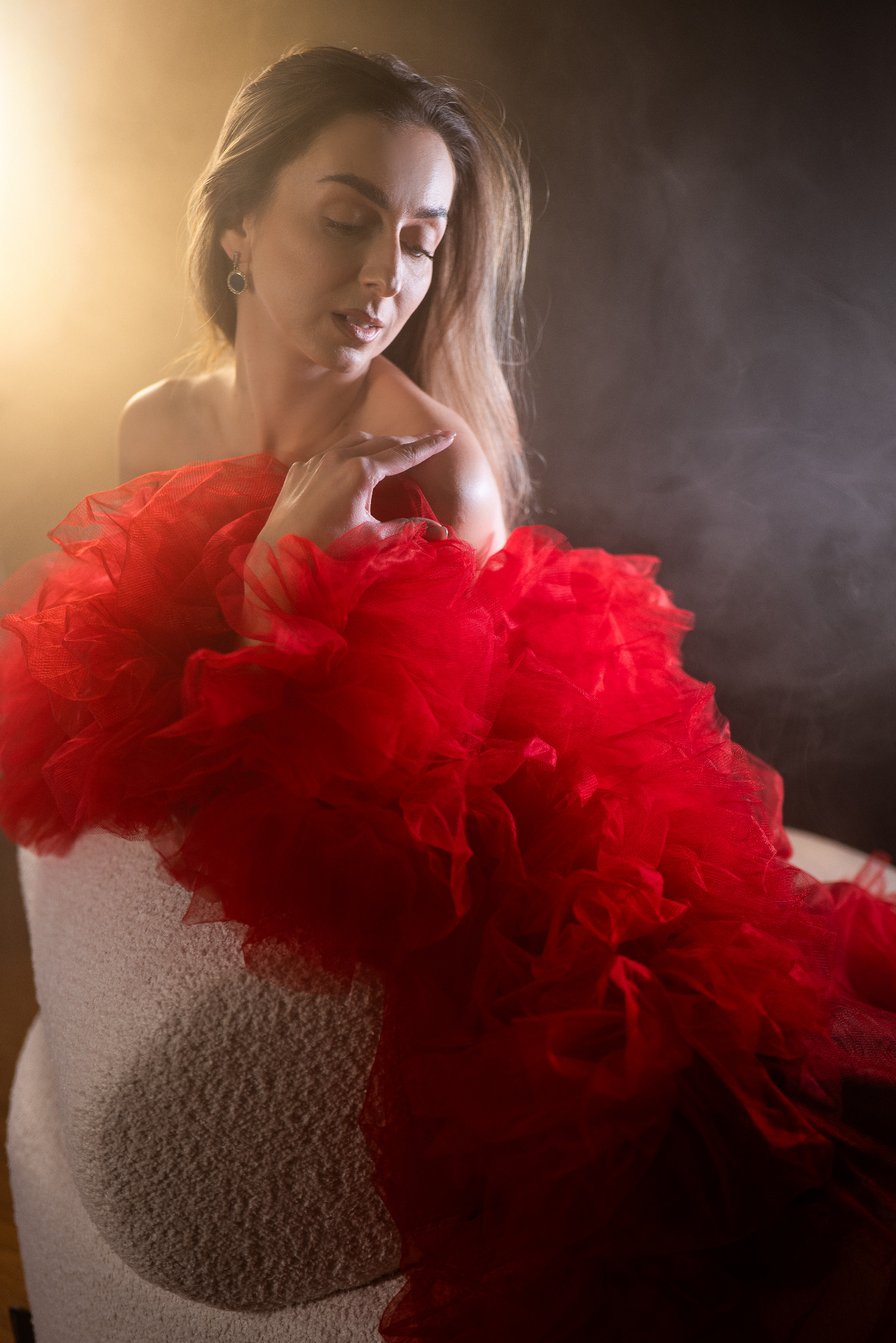 Portret kobiety w czerwonej, tiulowej sukni na jasnym fotelu, z delikatnym oświetleniem i ciemnym tłem, tworzący intymną i artystyczną atmosferę.