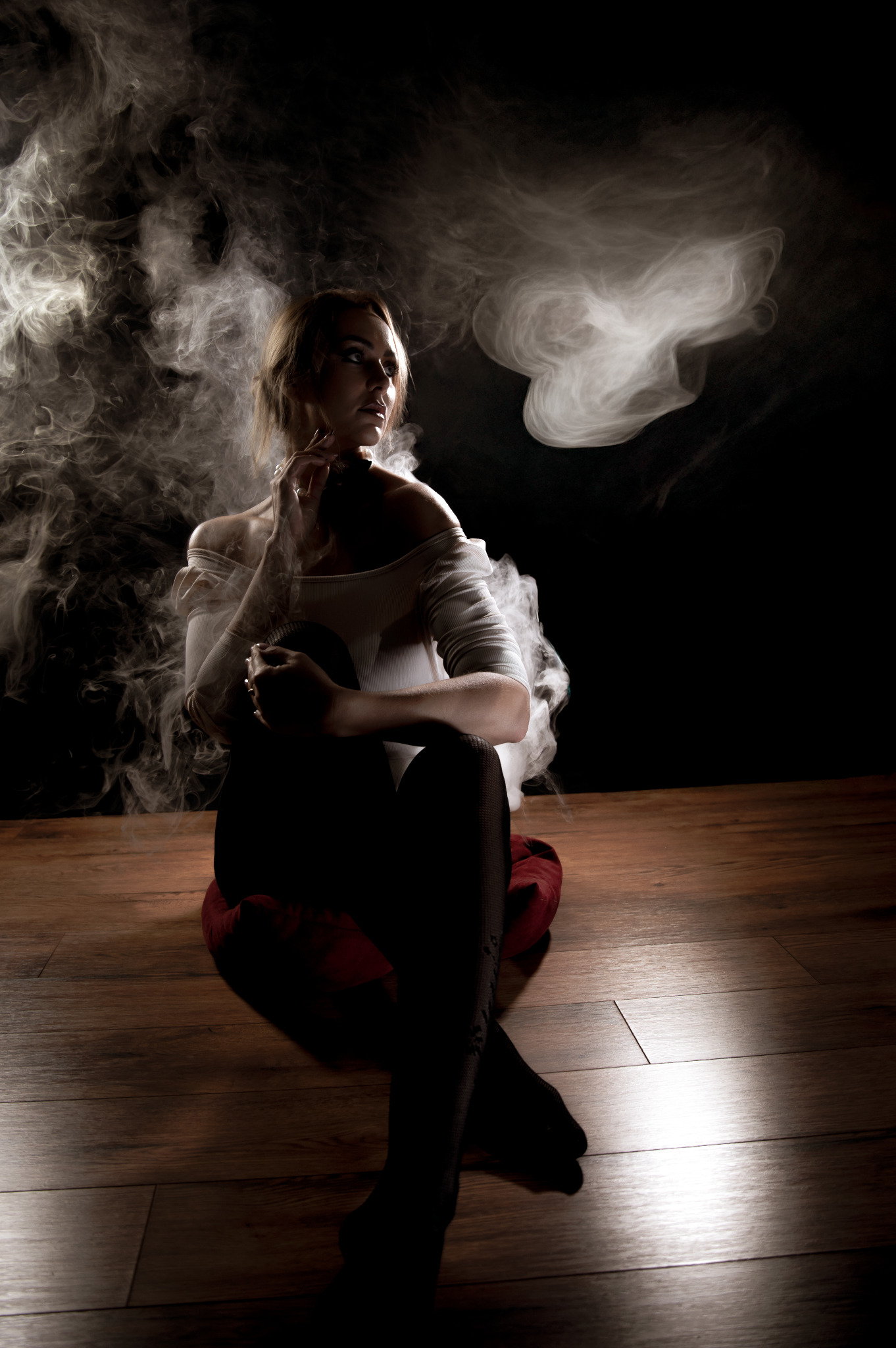 Kobieta siedząca na podłodze w otoczeniu dymu, w białym topie i czarnych rajstopach, zamyślona, w ciemnym otoczeniu. Artystyczne oświetlenie podkreśla kontury.