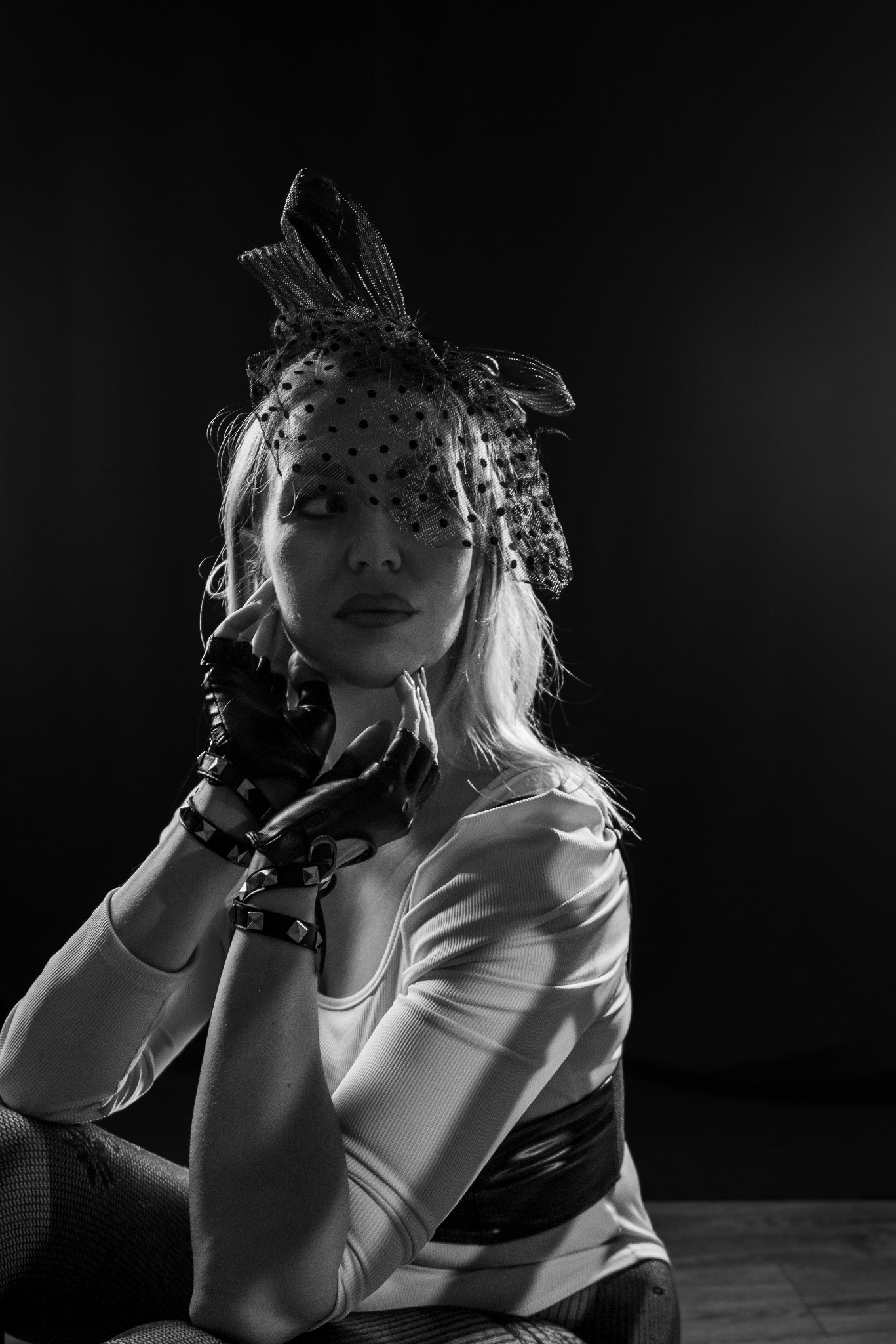 Czarno-białe zdjęcie kobiety w stylizowanej masce z siatki i skórzanych rękawicach z ćwiekami, pozującej na ciemnym tle. Artystyczny portret w klimacie retro.