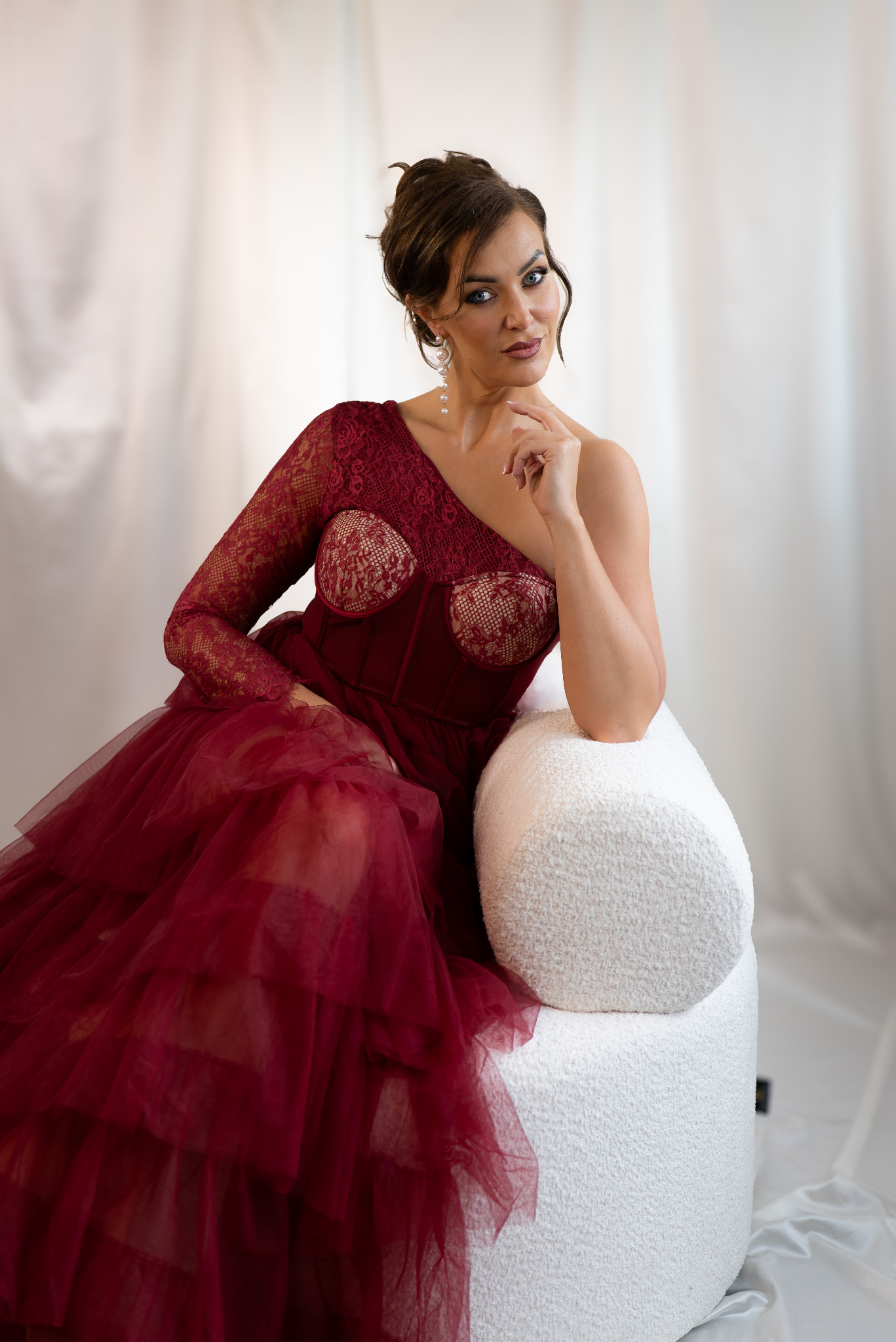 Kobieta w bordowej sukni z koronką, siedząca na białym fotelu. Uwagę przyciągają długie kolczyki i makijaż. Tło stanowi biała, delikatna tkanina.