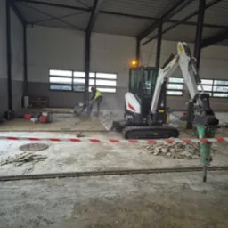 Prace rozbiórkowe w hali: minikoparka Bobcat E27z z wiertłem, pracownik z przecinarką do betonu. Widoczne gruz i taśma ostrzegawcza.
