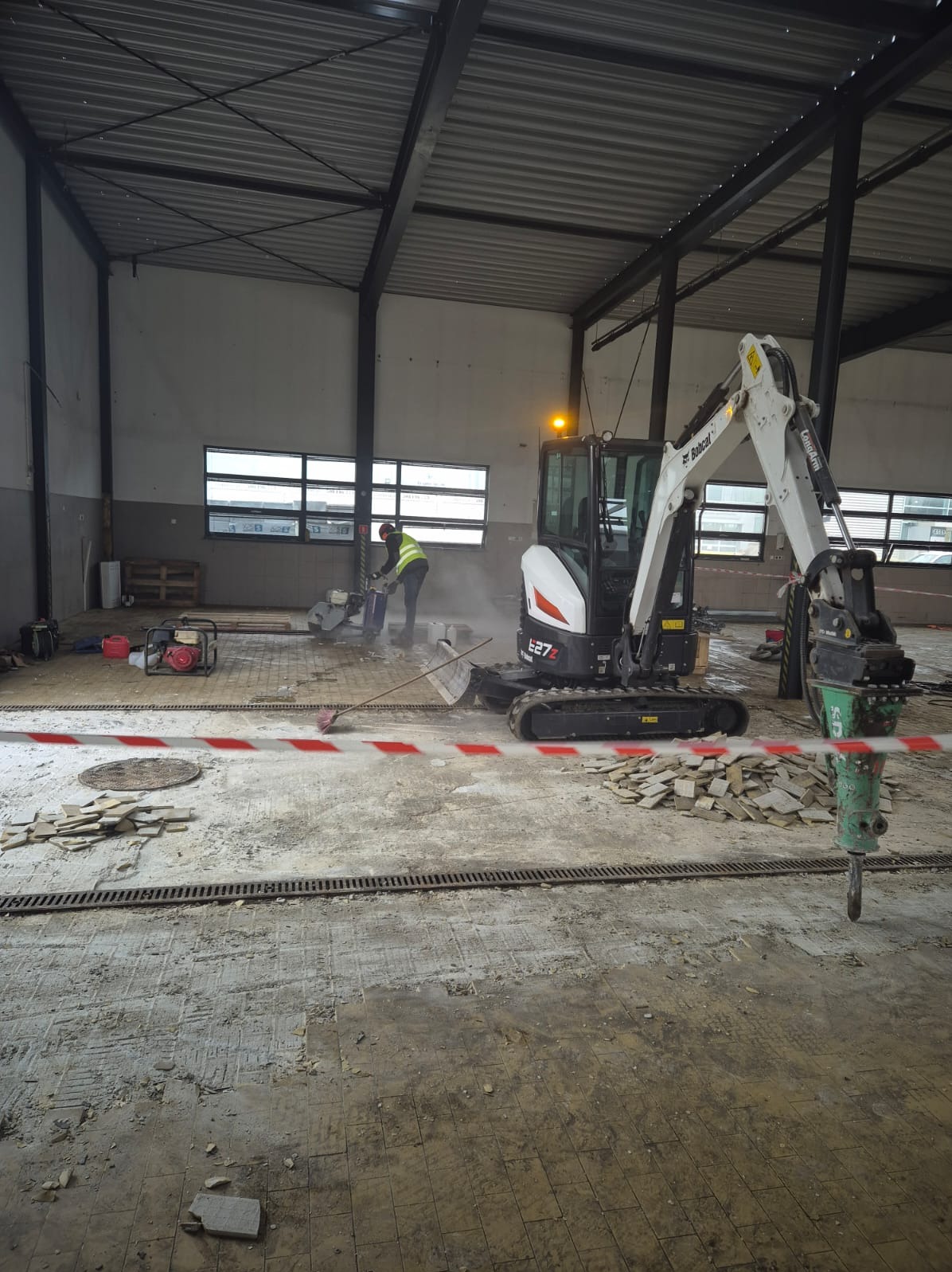 Prace rozbiórkowe w hali: minikoparka Bobcat E27z z wiertłem, pracownik z przecinarką do betonu. Widoczne gruz i taśma ostrzegawcza.