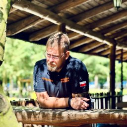 Akademia Reaguj i Ratuj - Instruktor z brodą i okularami, w koszulce z napisem INSTRUKTOR i flagą Polski, oparty o drewniany stół pod zadaszeniem w plenerze.