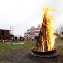 Akademia Reaguj i Ratuj - Ognisko w metalowej misie, wokół którego stoją ludzie, w tle budynki i drzewa, pochmurne niebo.