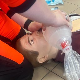 Akademia Reaguj i Ratuj - Instruktor w pomarańczowym stroju demonstruje resuscytację usta-usta na fantomie z użyciem worka samorozprężalnego. Widoczny ring na palcu instruktora i płytki podłogowe.